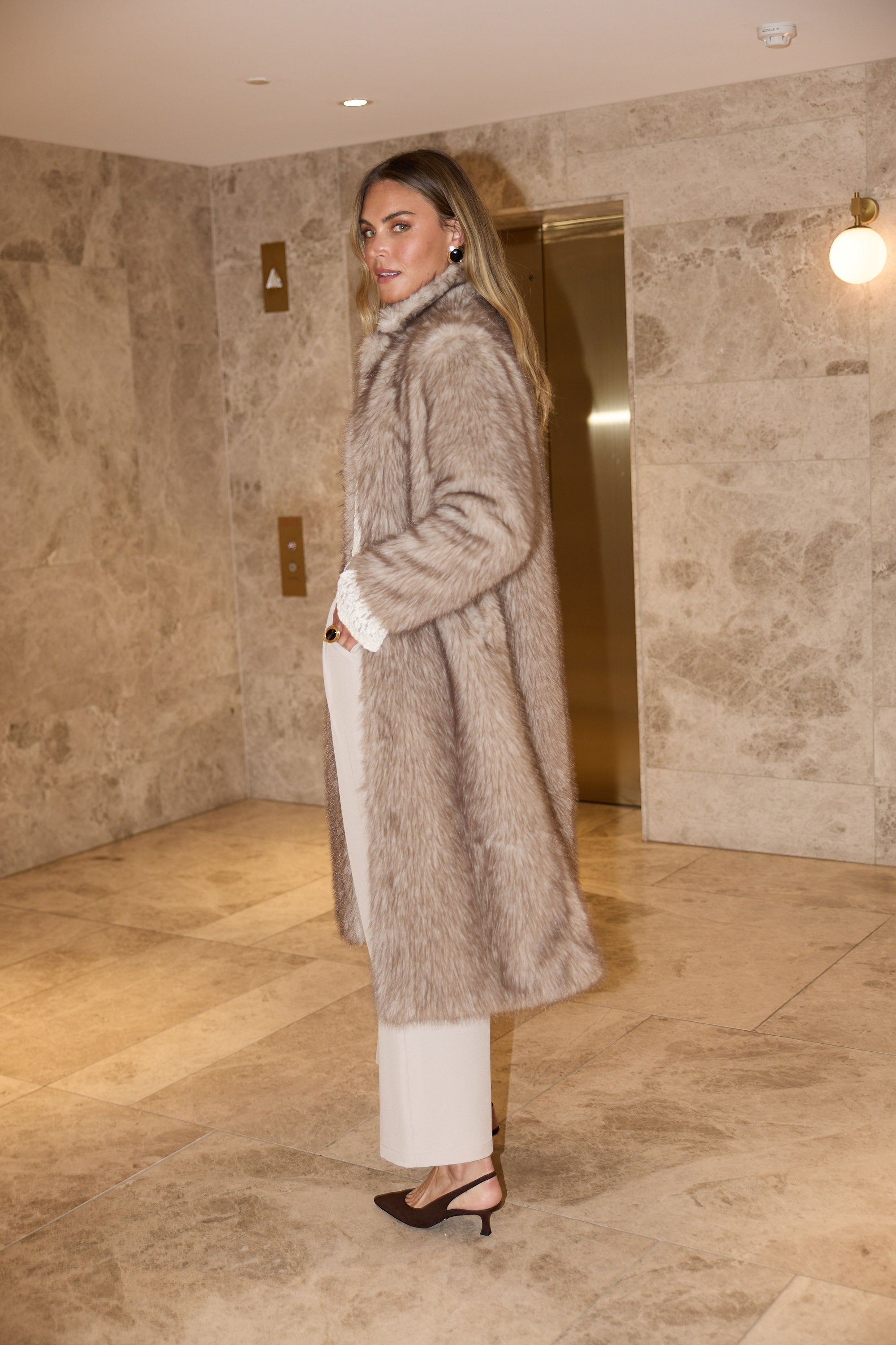 Woman wearing a fur coat in an indoor setting with stone walls and a light fixture.