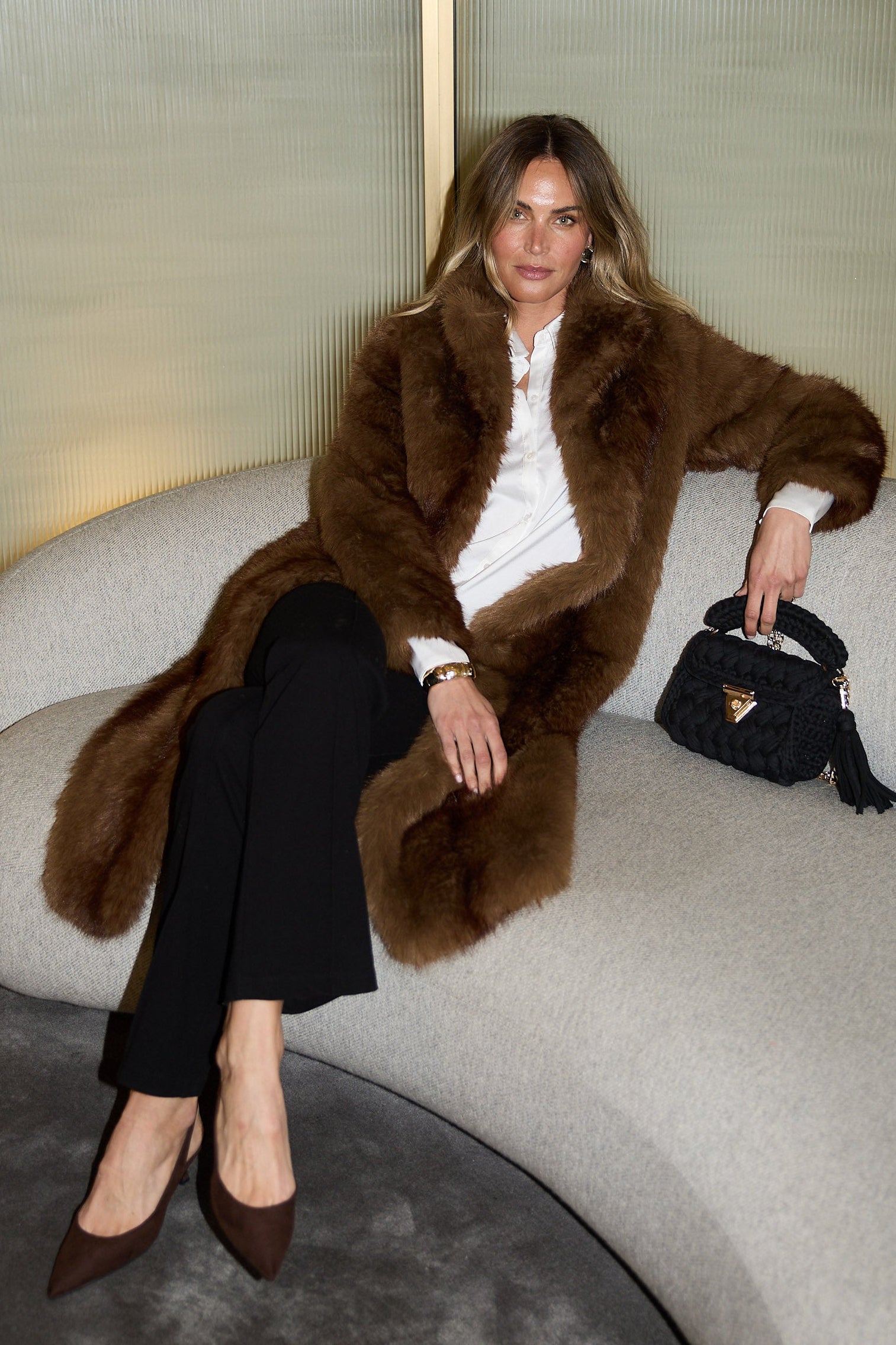 Woman in a fur coat sitting on a couch holding a handbag.