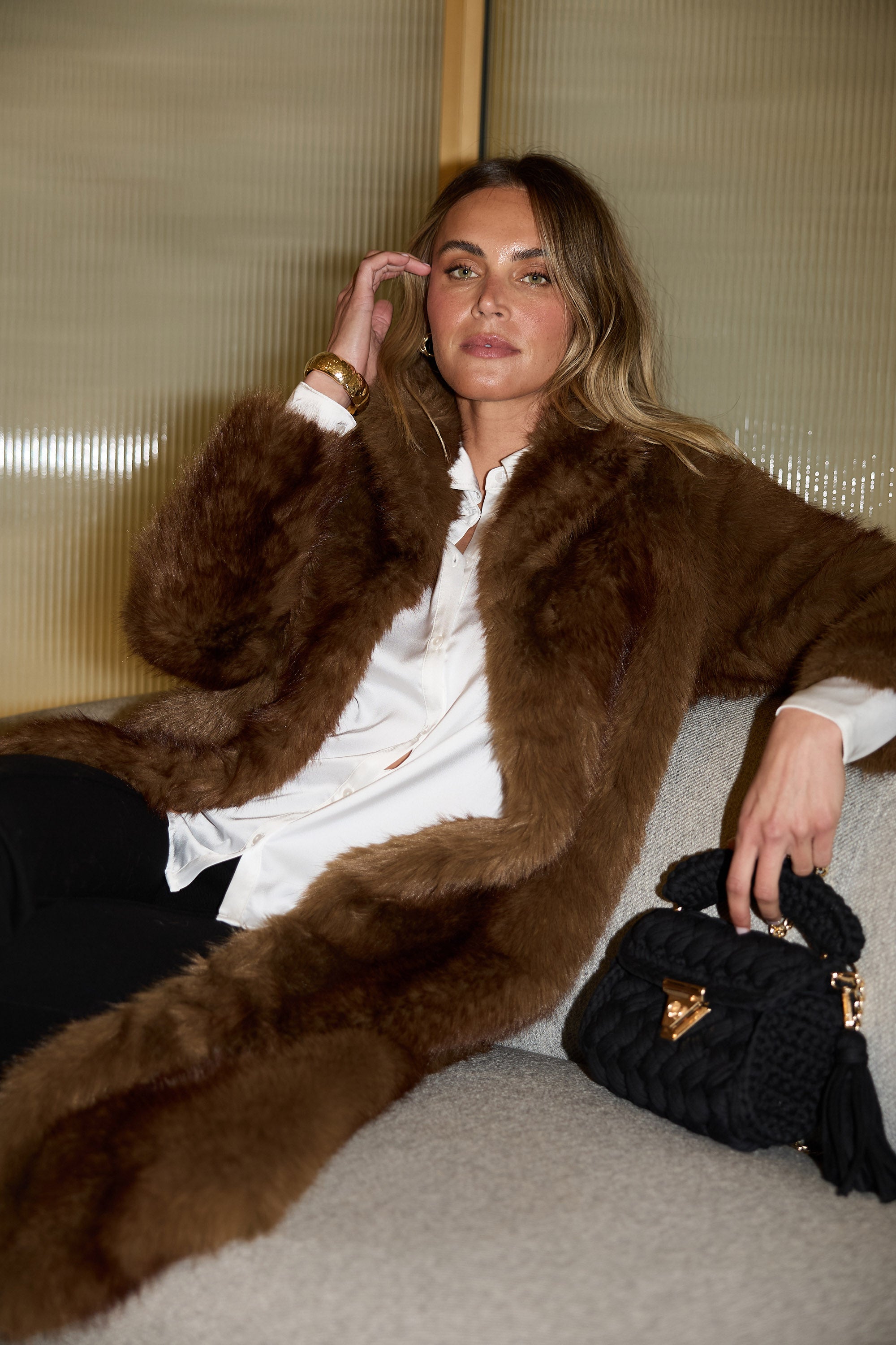 Woman wearing a brown fur coat sitting on a couch with a black handbag.