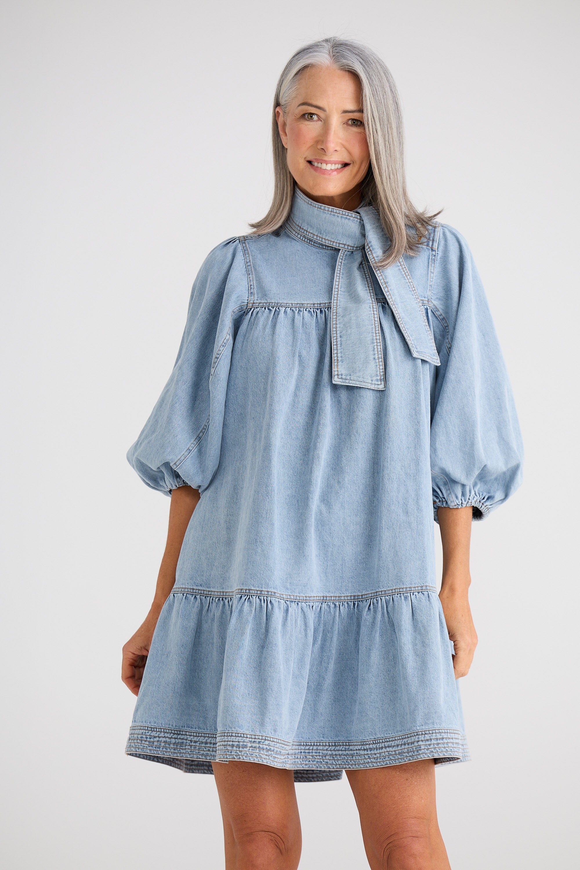 Woman wearing a light blue denim dress with a bow detail on a white background