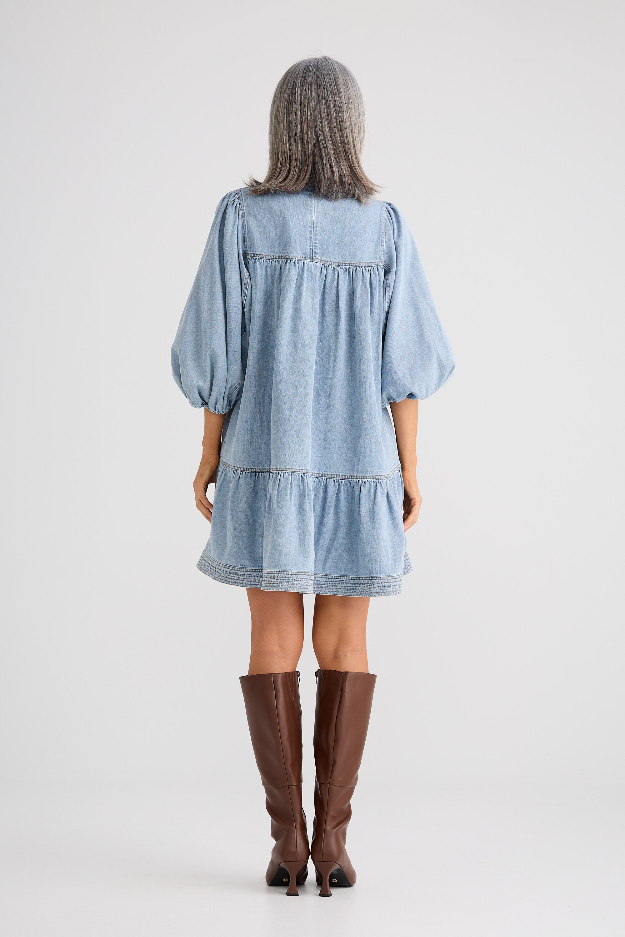 Woman wearing a light blue denim dress with brown knee-high boots on a white background