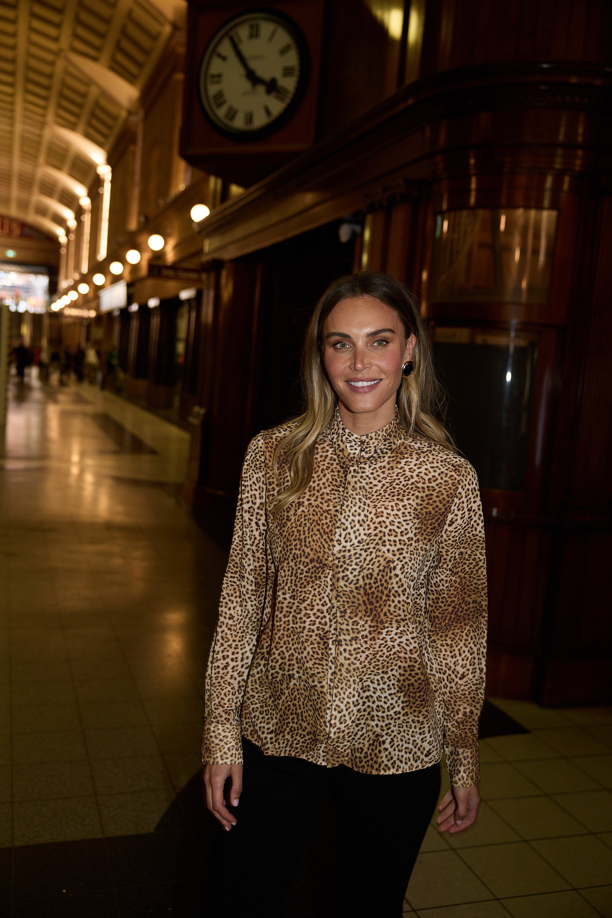 Woman wearing a leopard print blouse in a large, dimly lit hall with a clock on the wall.
