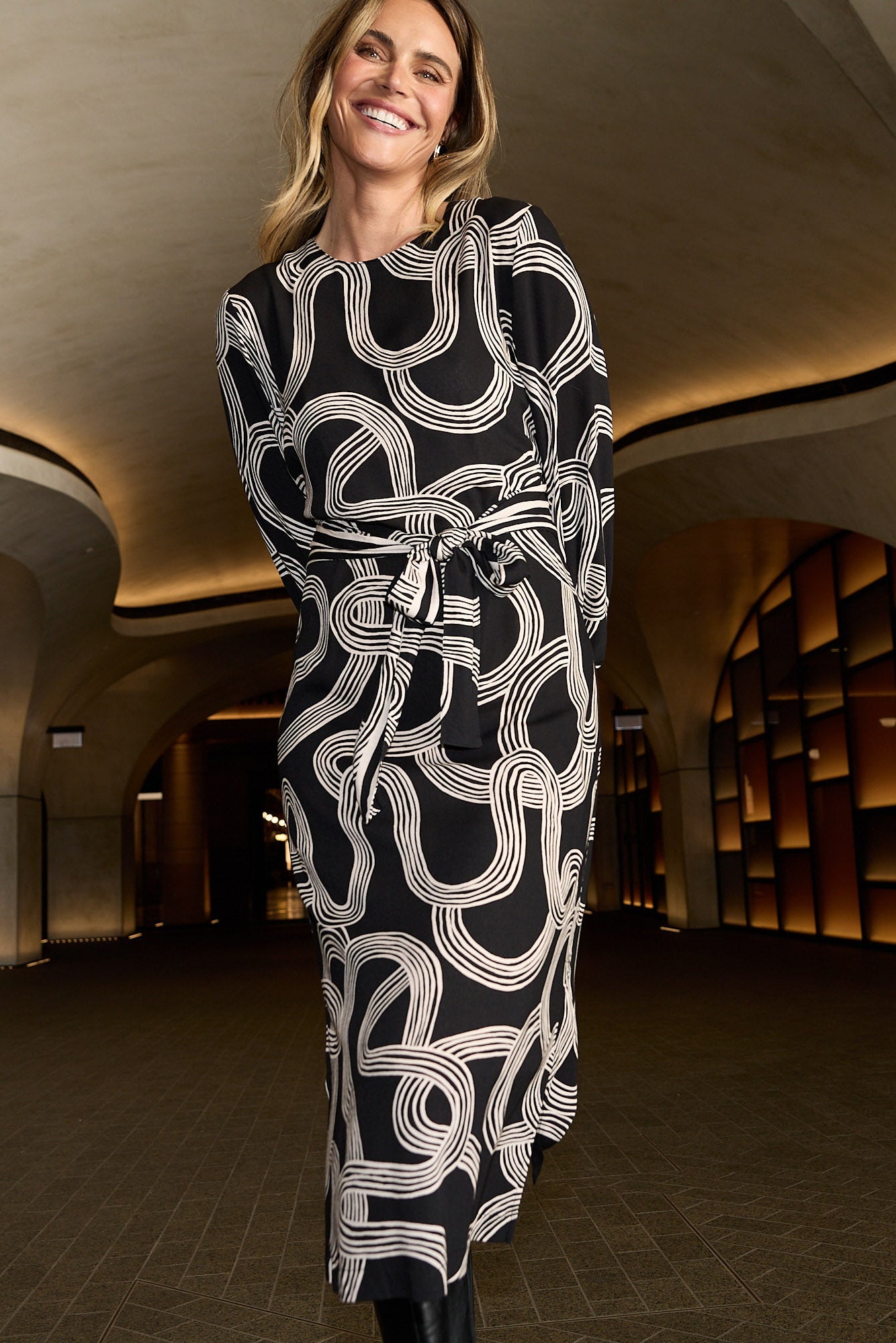 Woman in a black and white patterned coat walking through an architectural interior.
