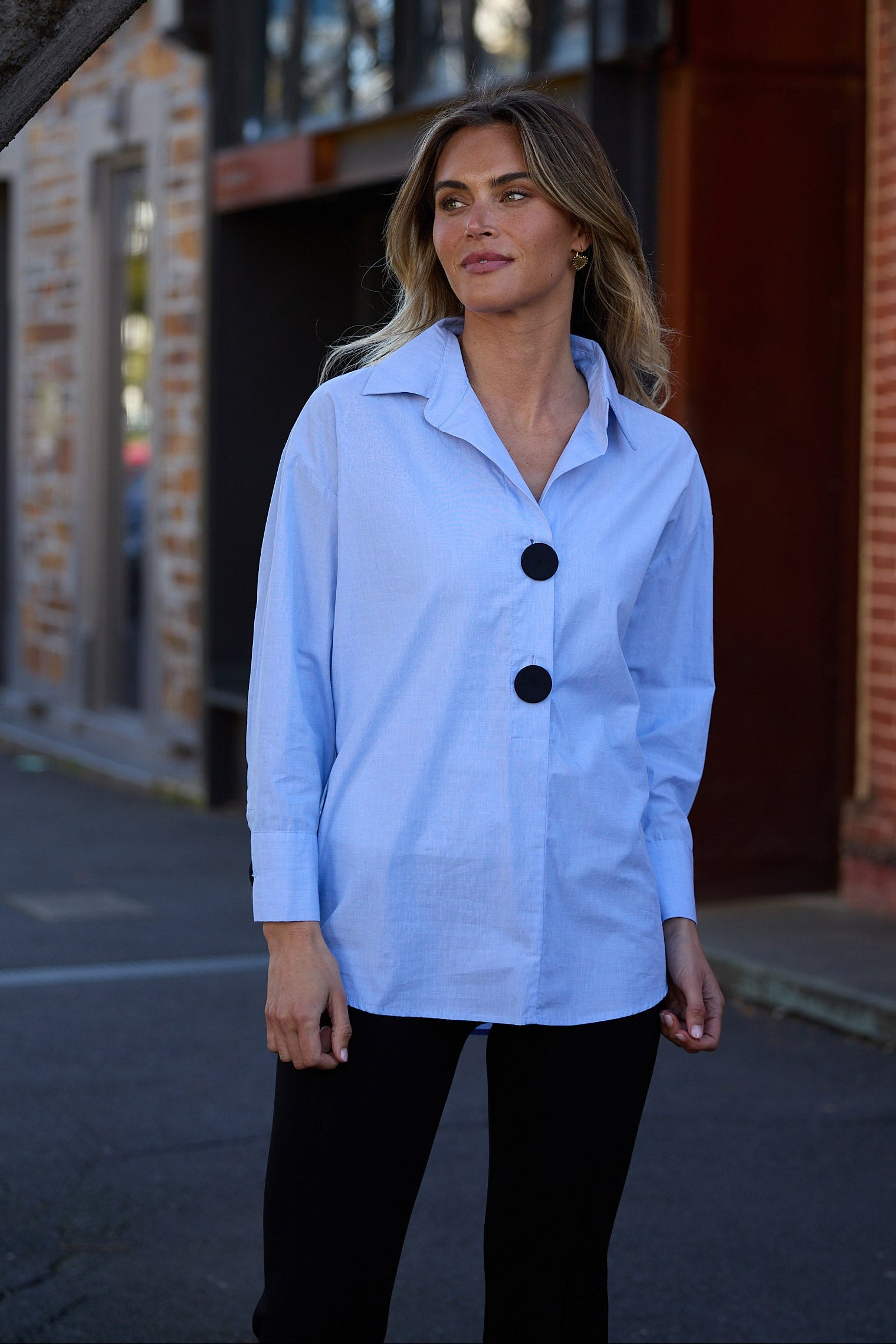 Woman wearing a light blue jacket with black buttons standing in front of a brick building.