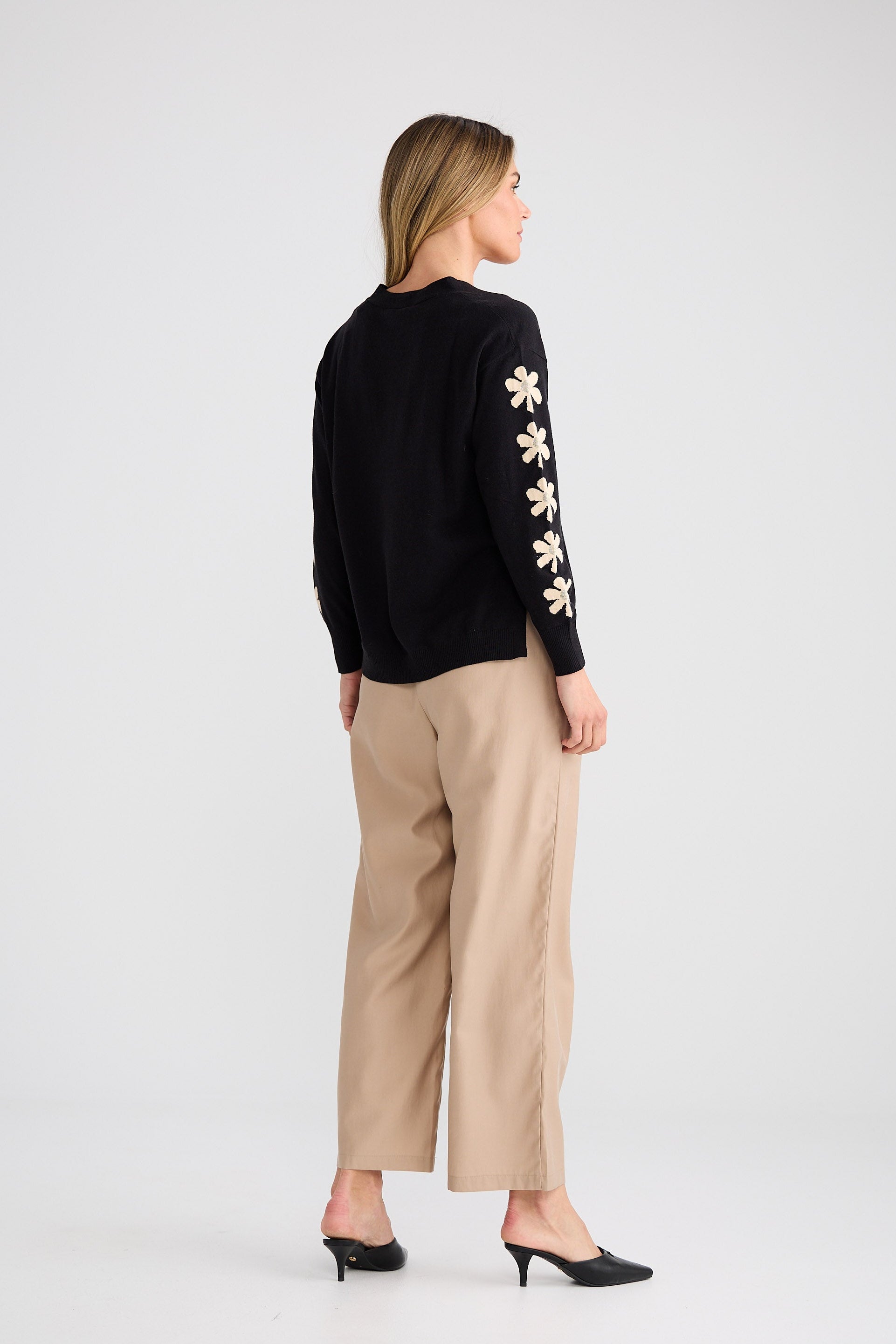 Woman wearing a black sweater with floral designs on the sleeves and beige pants against a white background