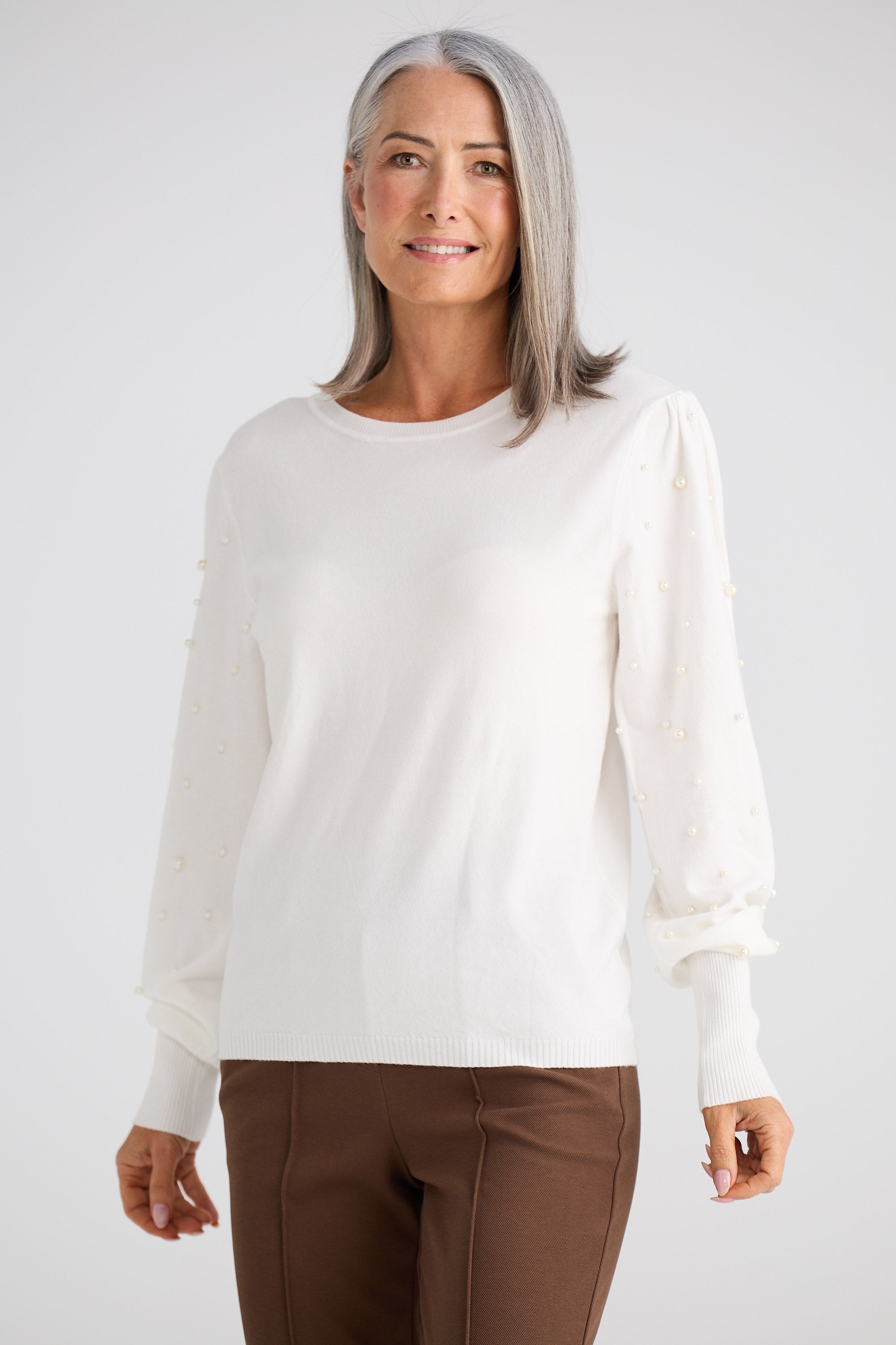 Woman wearing a white blouse and brown pants against a plain background