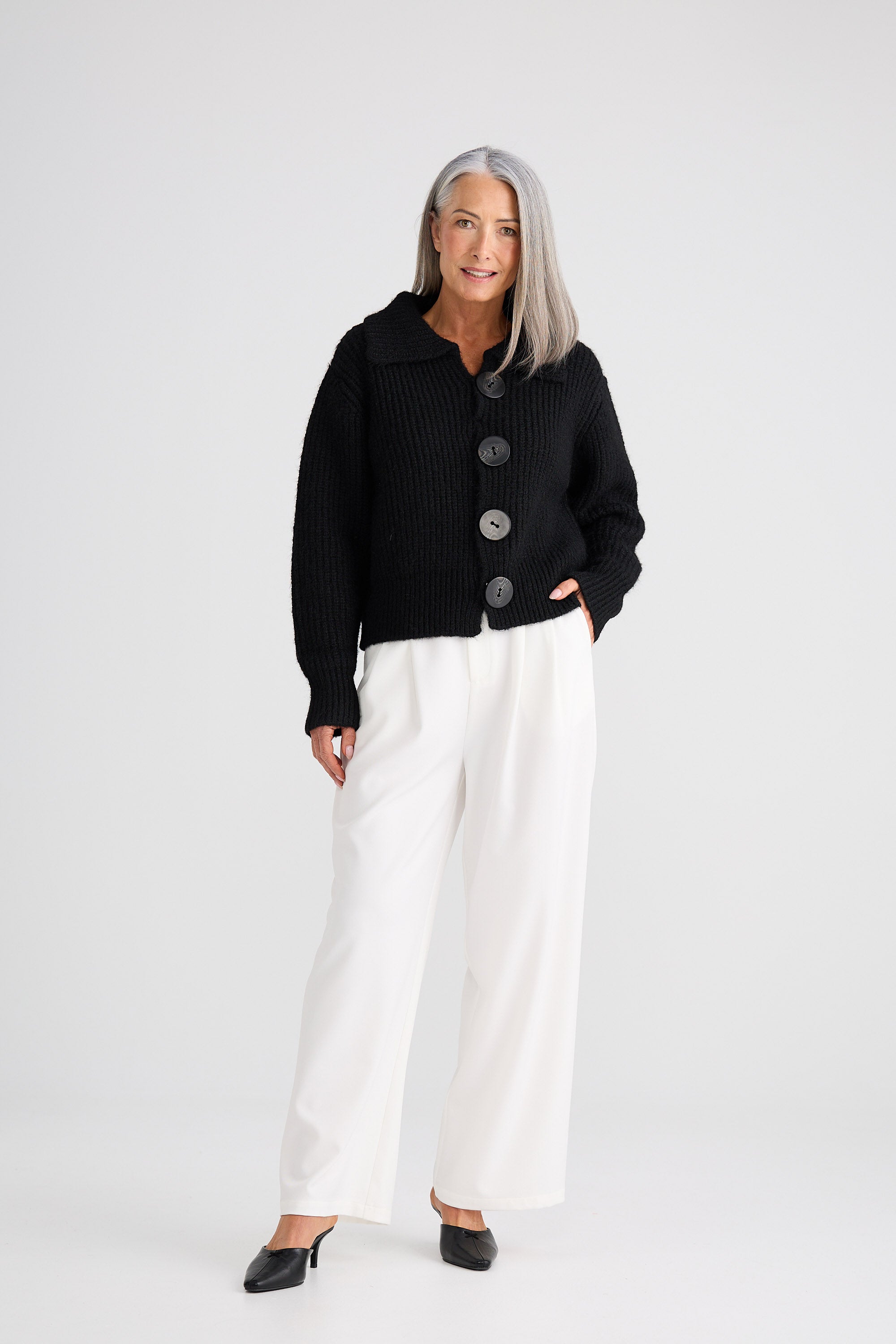Woman wearing a black cardigan with buttons and white pants on a white background
