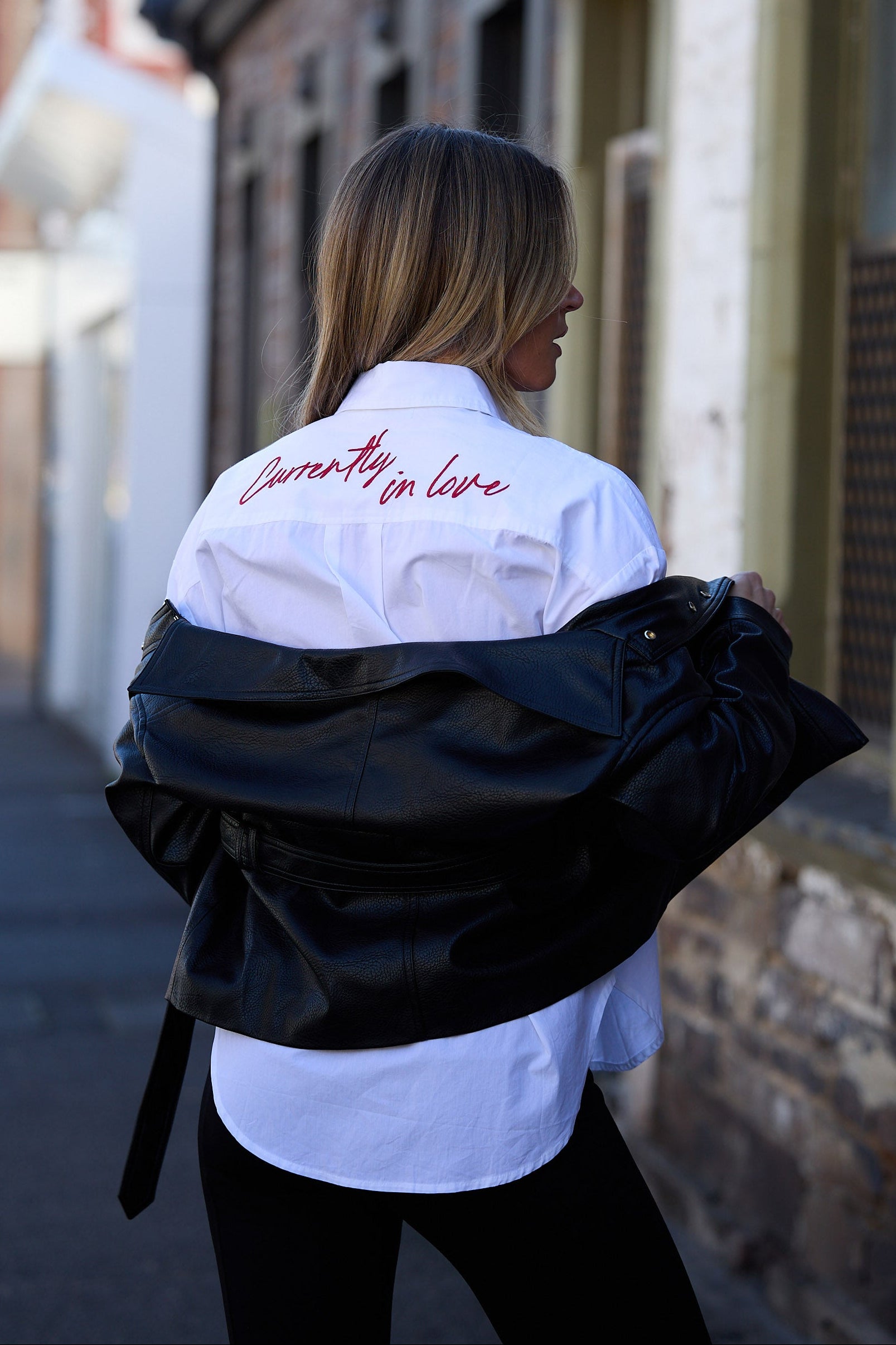 Person wearing a black jacket over a white shirt with text, standing on a street.