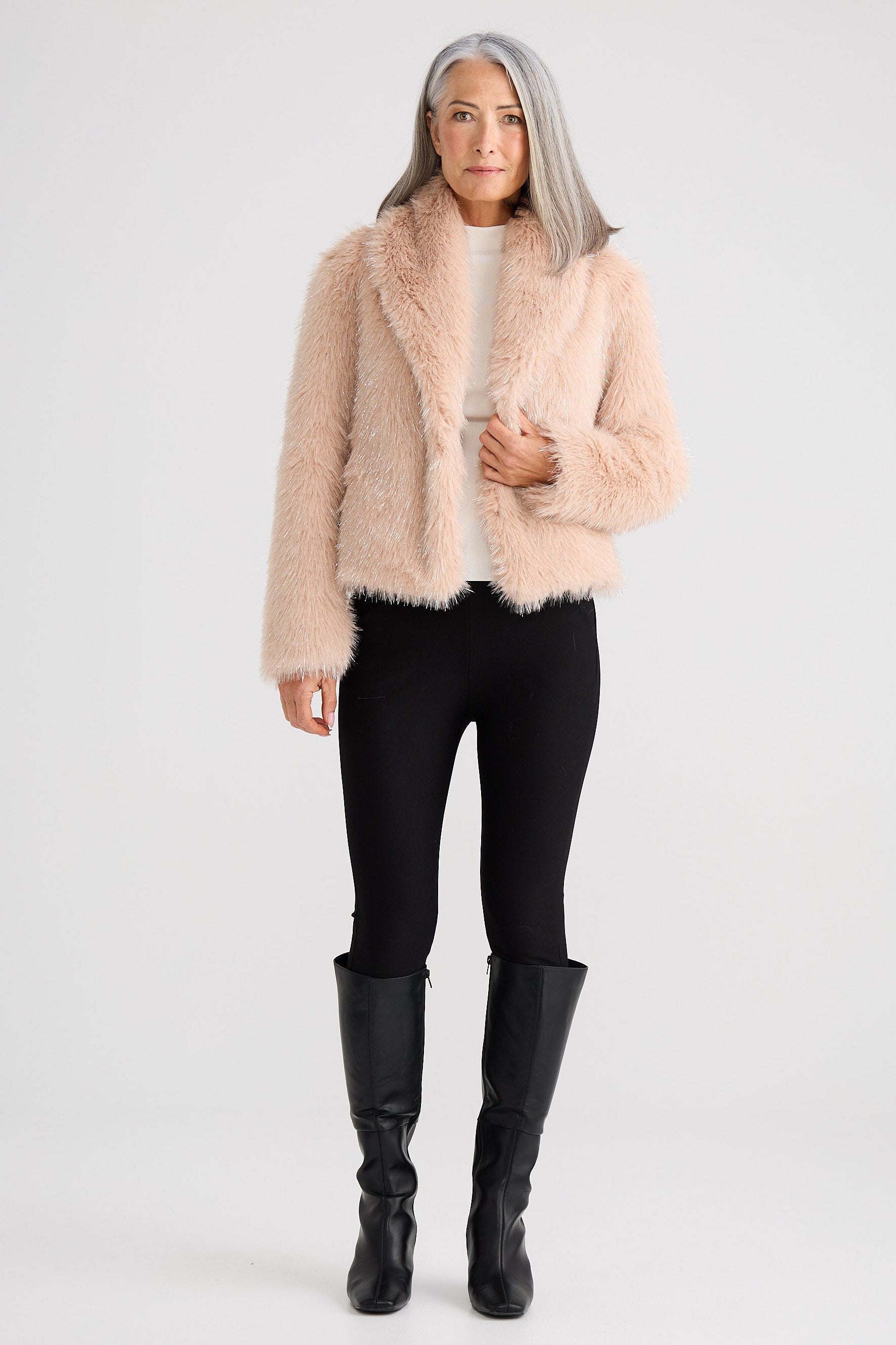 Woman wearing a beige faux fur jacket, black leggings, and black boots on a white background