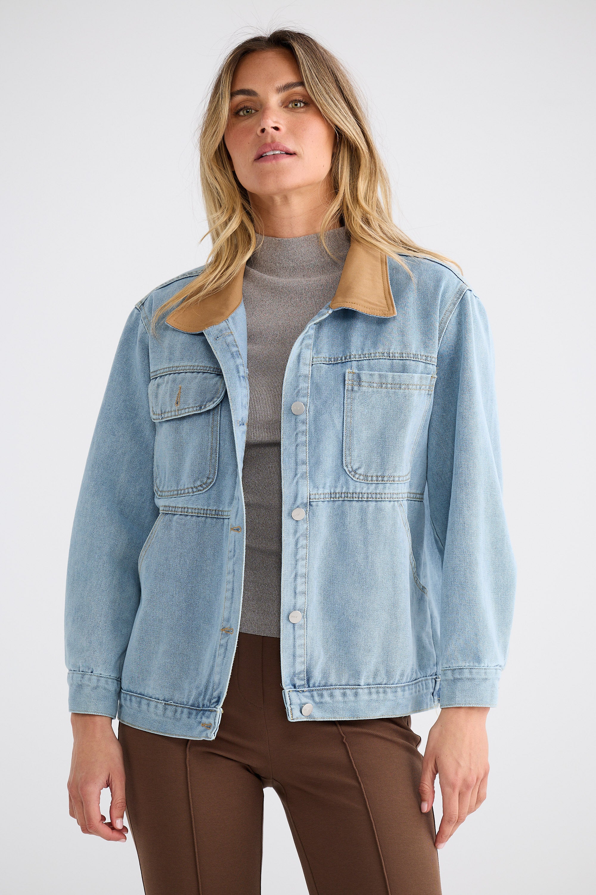 Woman wearing a light blue denim jacket over a gray top and brown pants on a white background