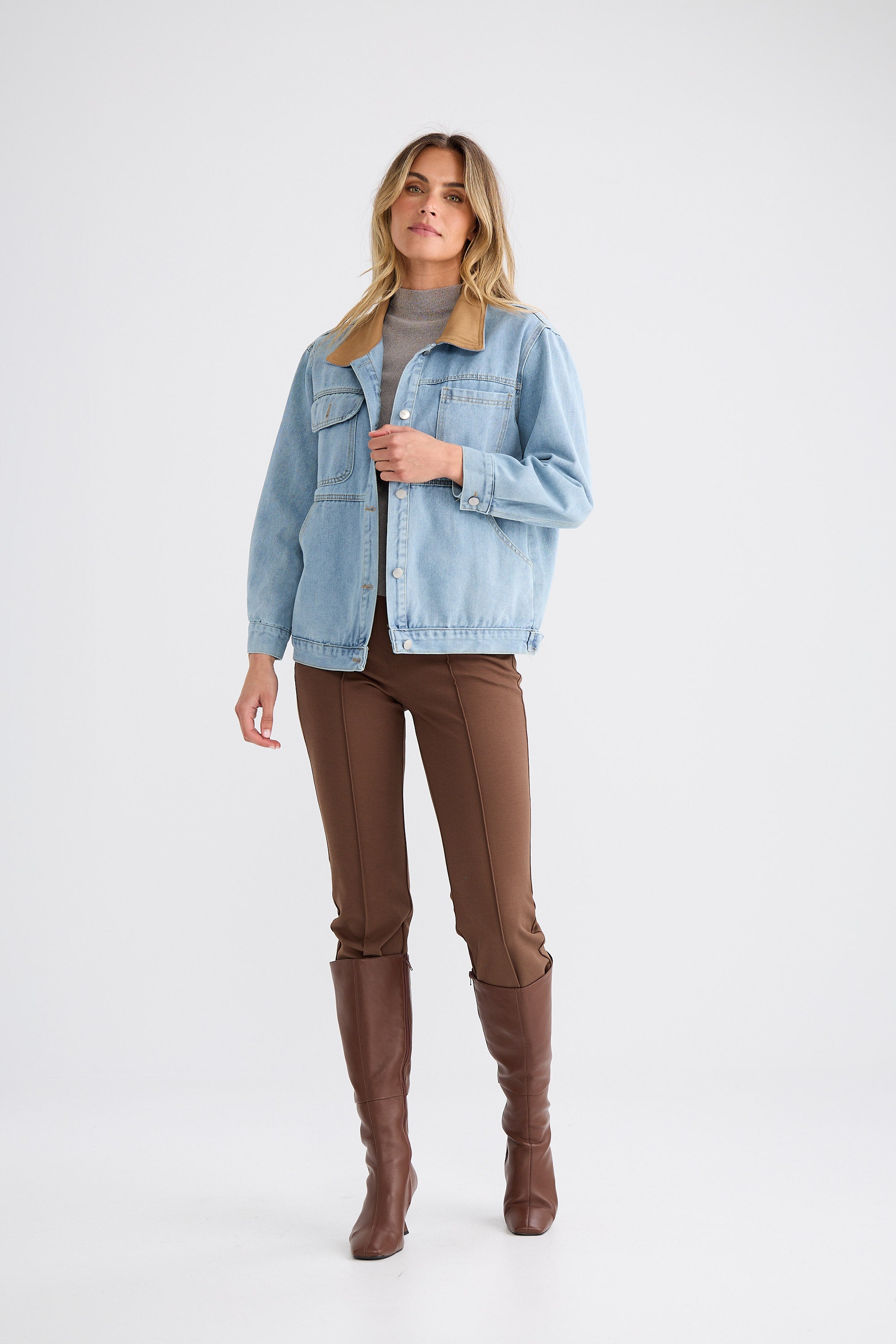 Woman wearing a light blue denim jacket, brown pants, and brown knee-high boots on a white background