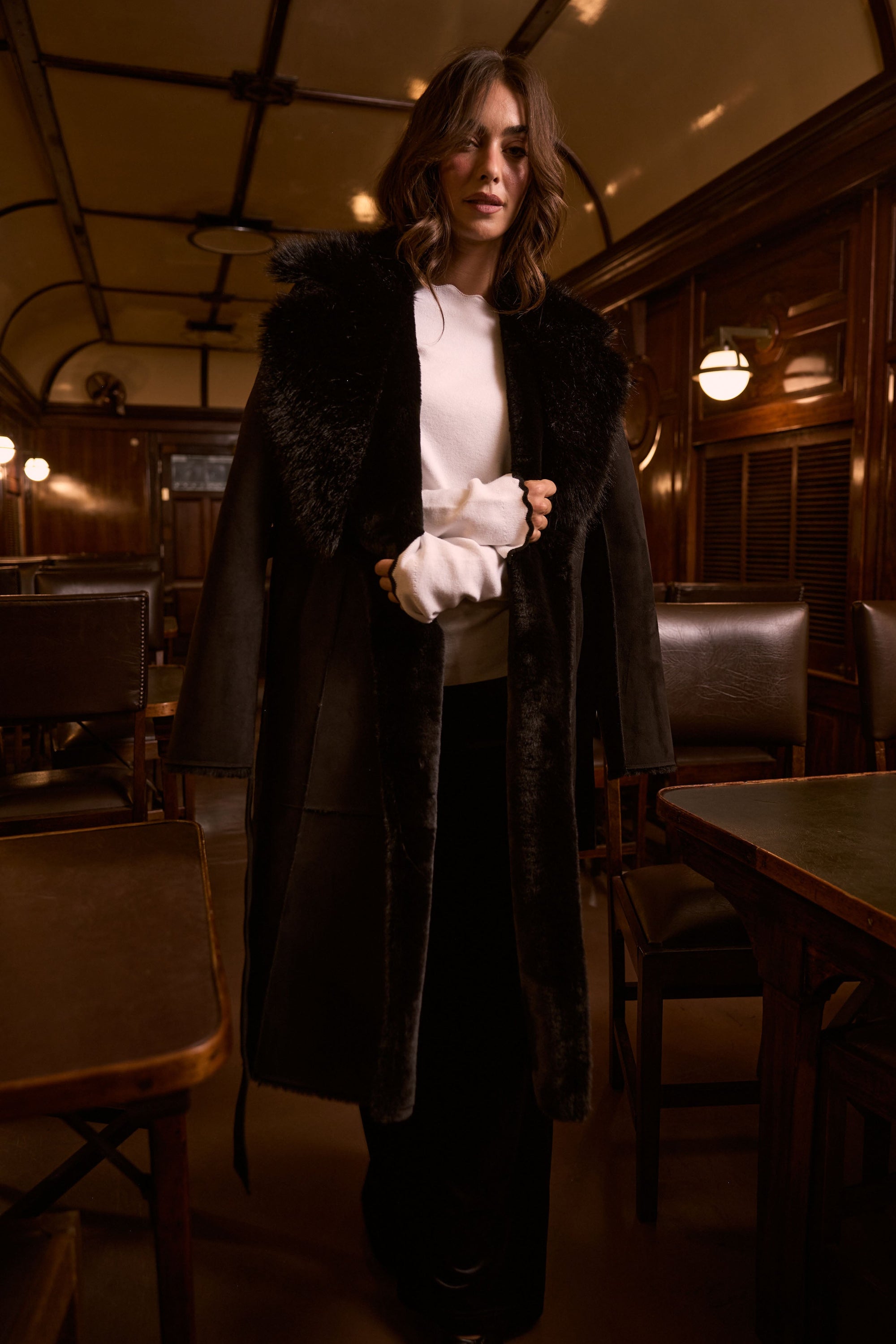 Model wearing Rosamund Fur Coat in black with notched lapels and waist tie, side view on white background