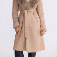 Model wearing Rosamund Fur Coat in beige with notched lapels and waist tie, side view on white background