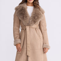 Model wearing Rosamund Fur Coat in beige with notched lapels and waist tie, side view on white background