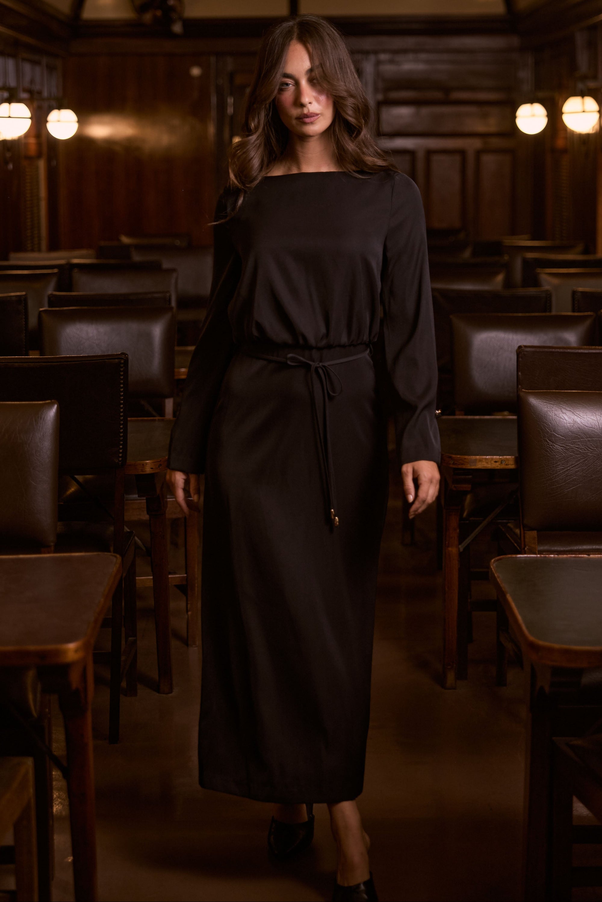 Model wearing Kensington Dress in black with boat neckline and long sleeves, side view on white background