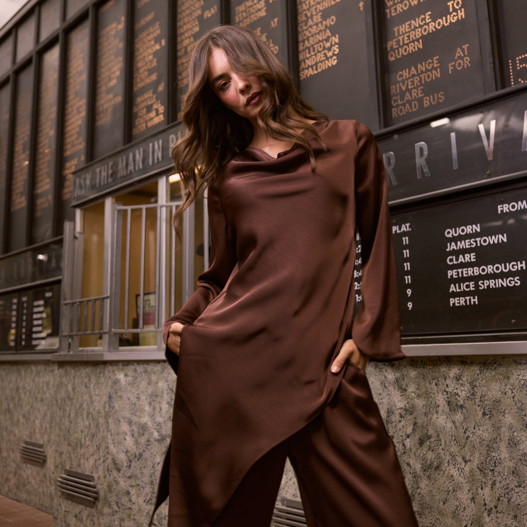 Woman in a brown outfit standing in front of a train departure board.