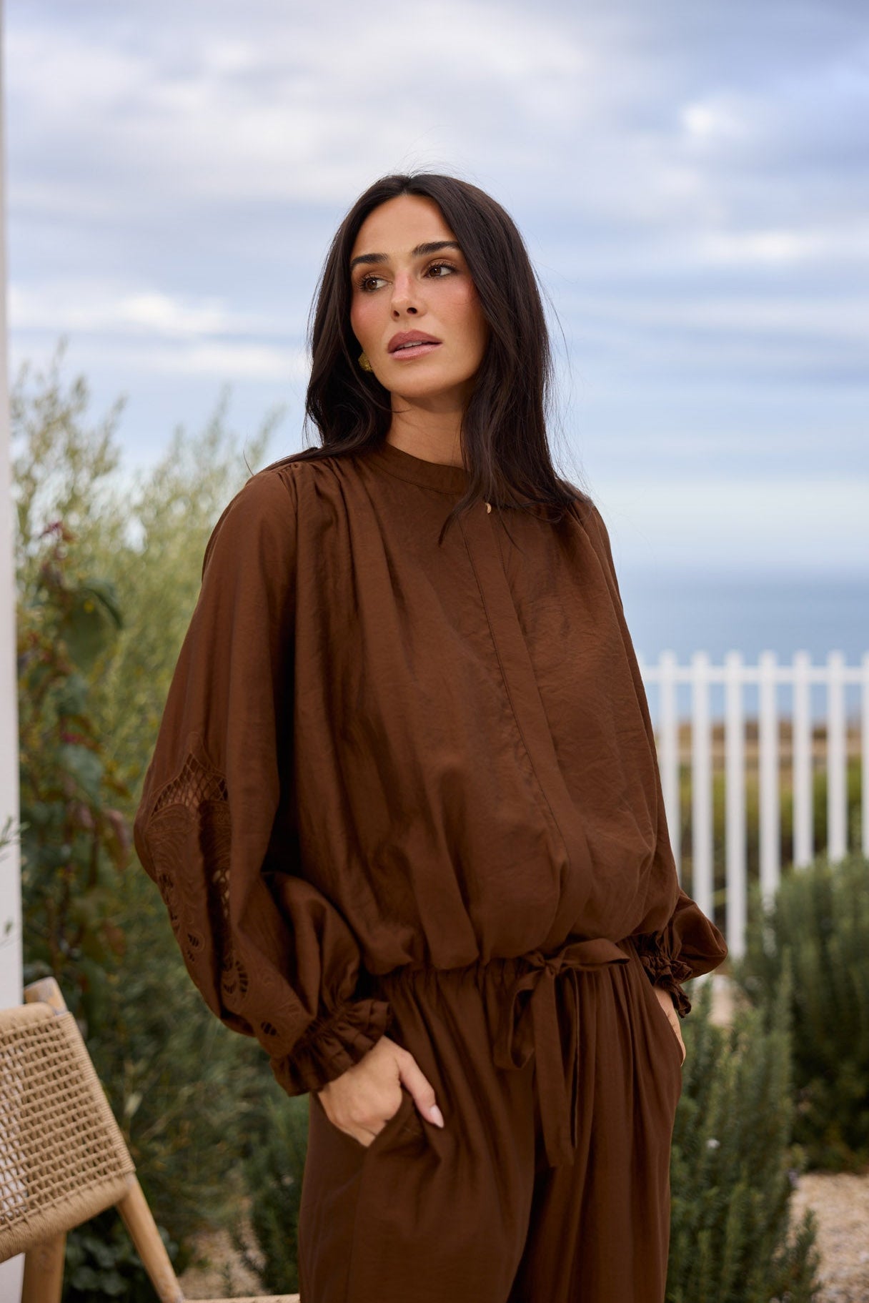 A woman wearing a brown long-sleeve jumpsuit with a tie waist, standing outdoors in front of a white fence and sky.