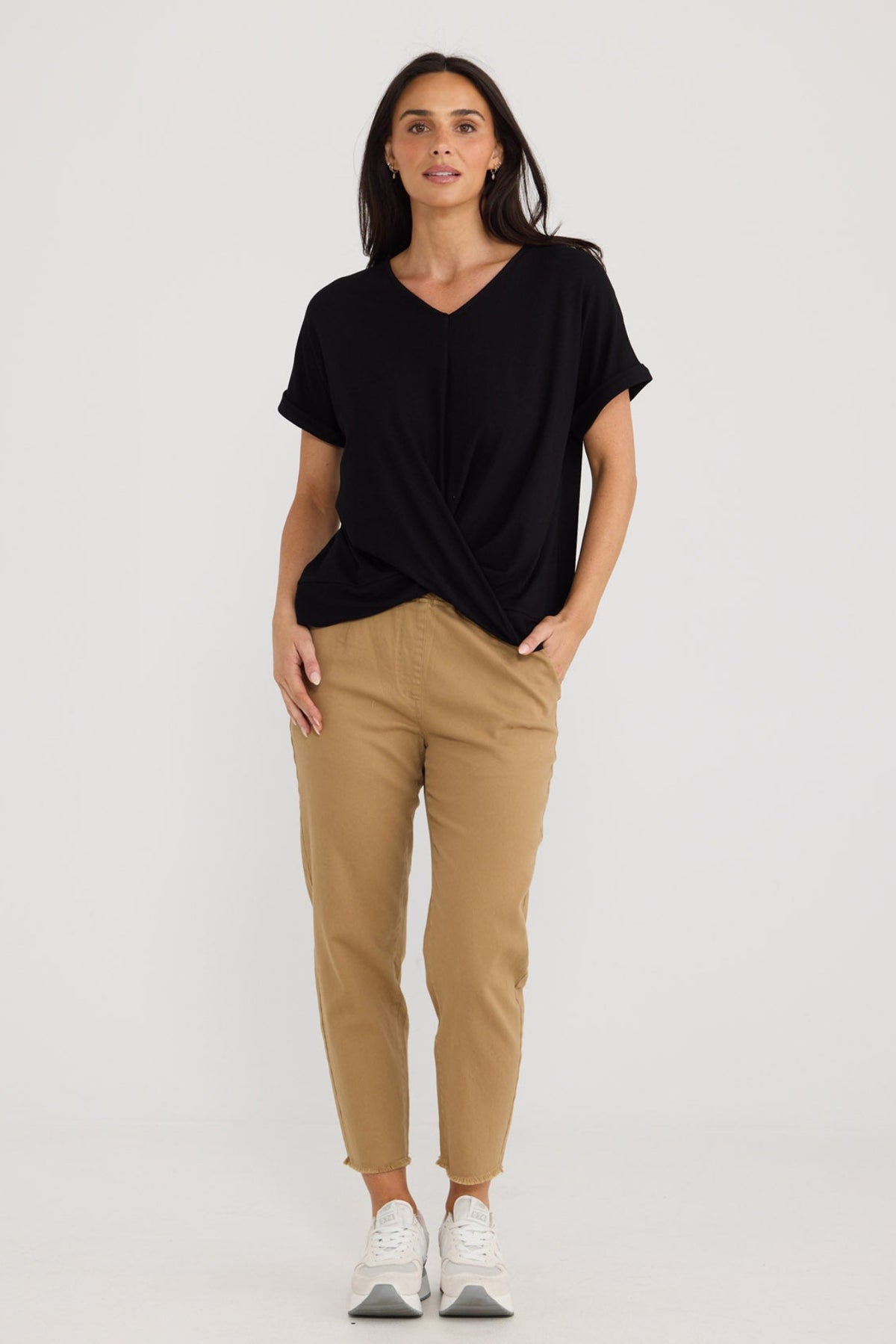 A woman modeling a black short-sleeve V-neck top and tan cropped trousers with white sneakers, standing against a plain light gray studio background by Brave + True.