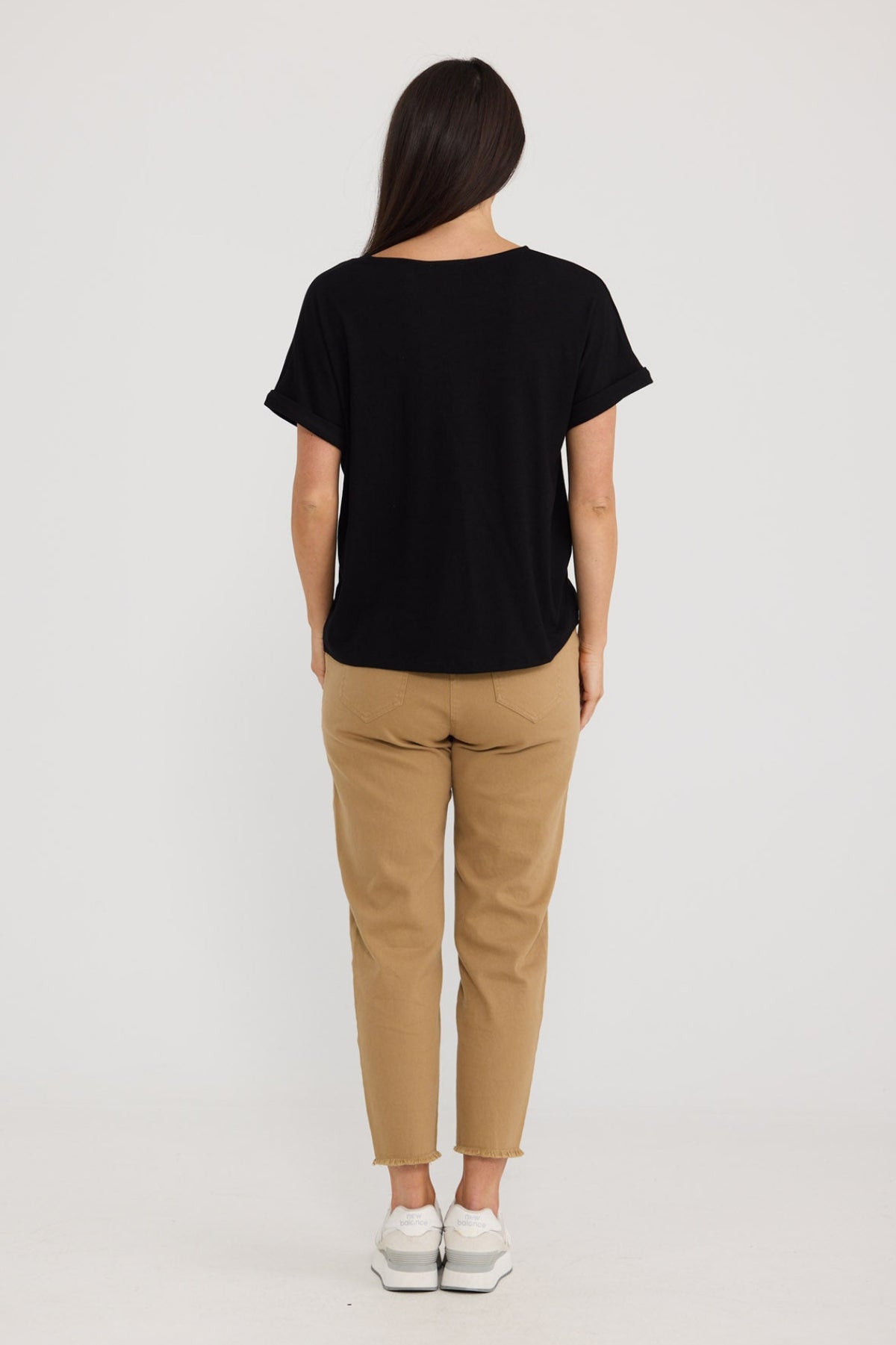 A woman viewed from behind wearing a black short-sleeve t-shirt, tan cropped pants, and white sneakers against a plain light gray studio background by Brave + True.
