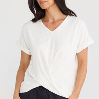 A woman wearing a white short-sleeve V-neck top with a twisted front detail, paired with dark pants, against a light gray studio background by Brave + True.