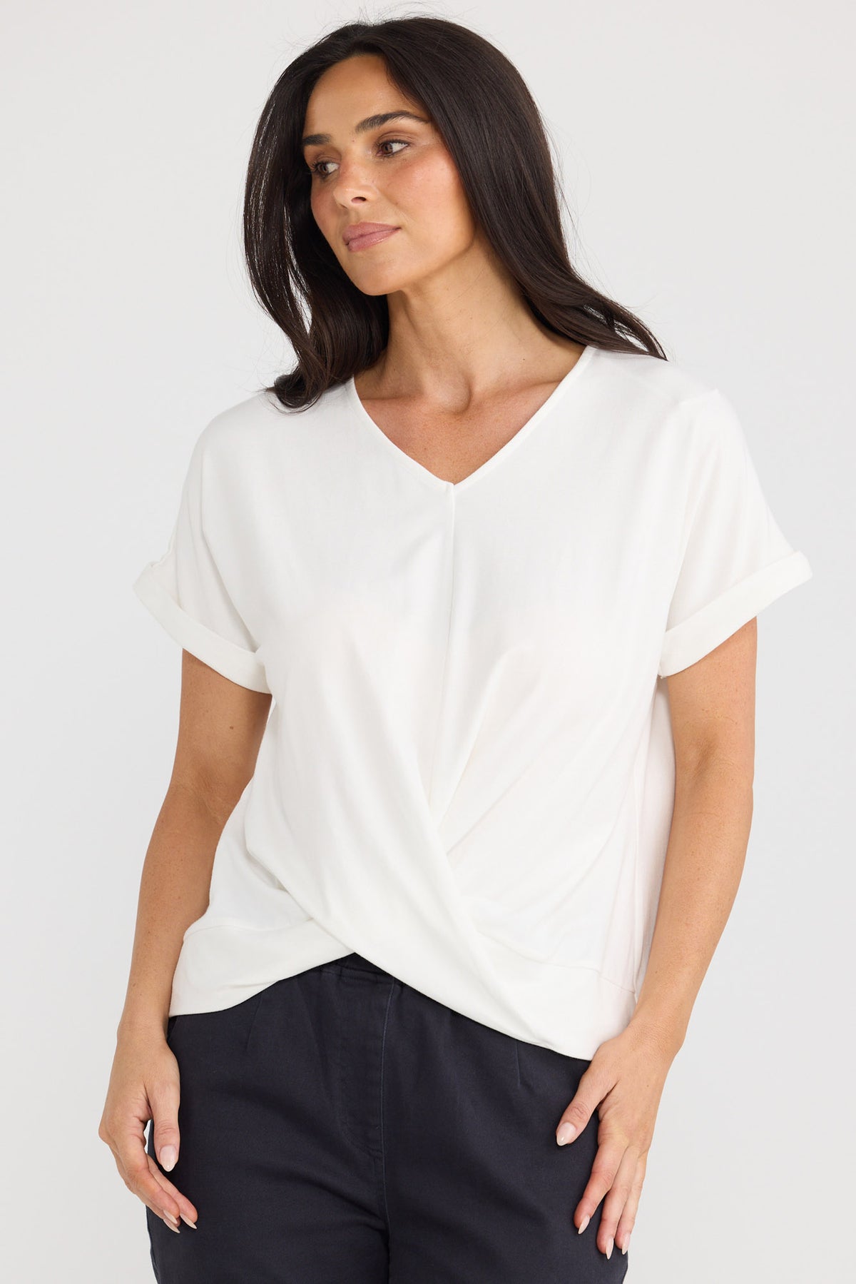 A woman wearing a white short-sleeve V-neck top with a twisted front detail, paired with dark pants, against a light gray studio background by Brave + True.
