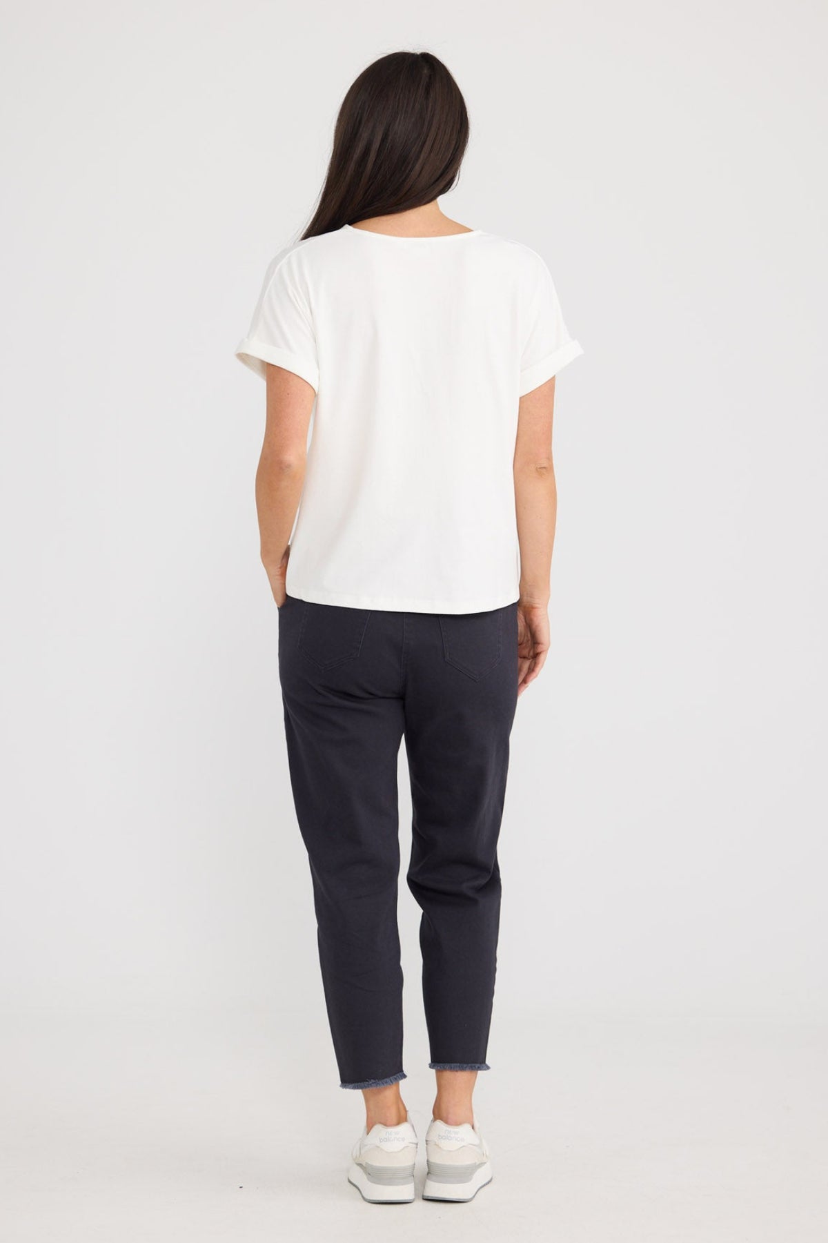 A woman viewed from behind wearing a white short-sleeve t-shirt, dark navy pants, and white sneakers, standing against a plain white studio background by Brave + True.