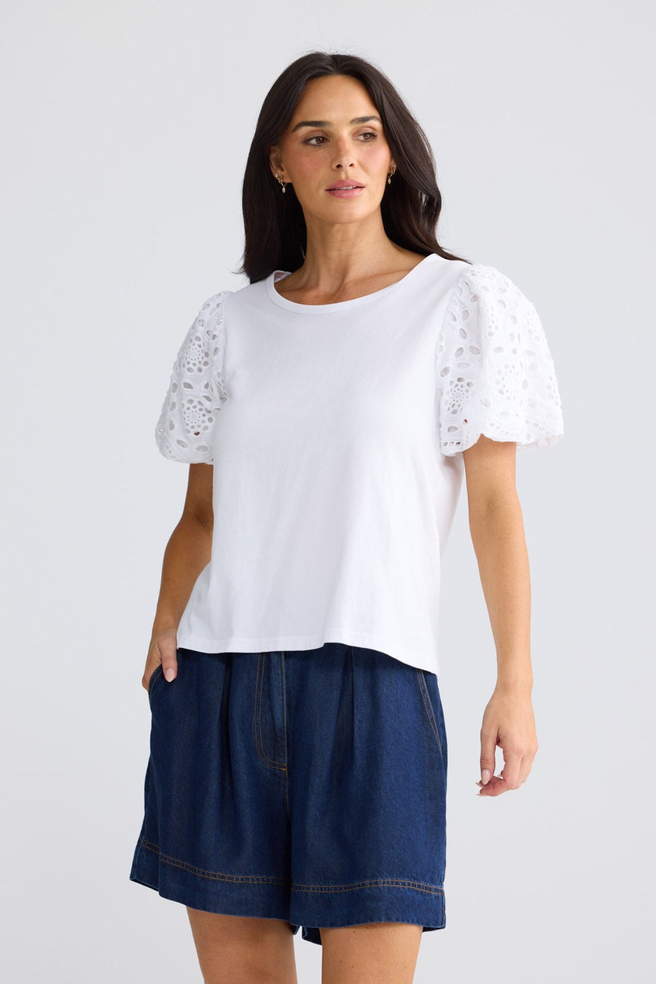 A woman modeling a white short-sleeve top with eyelet lace puff sleeves, paired with dark blue denim shorts, against a light gray studio background by Brave + True.