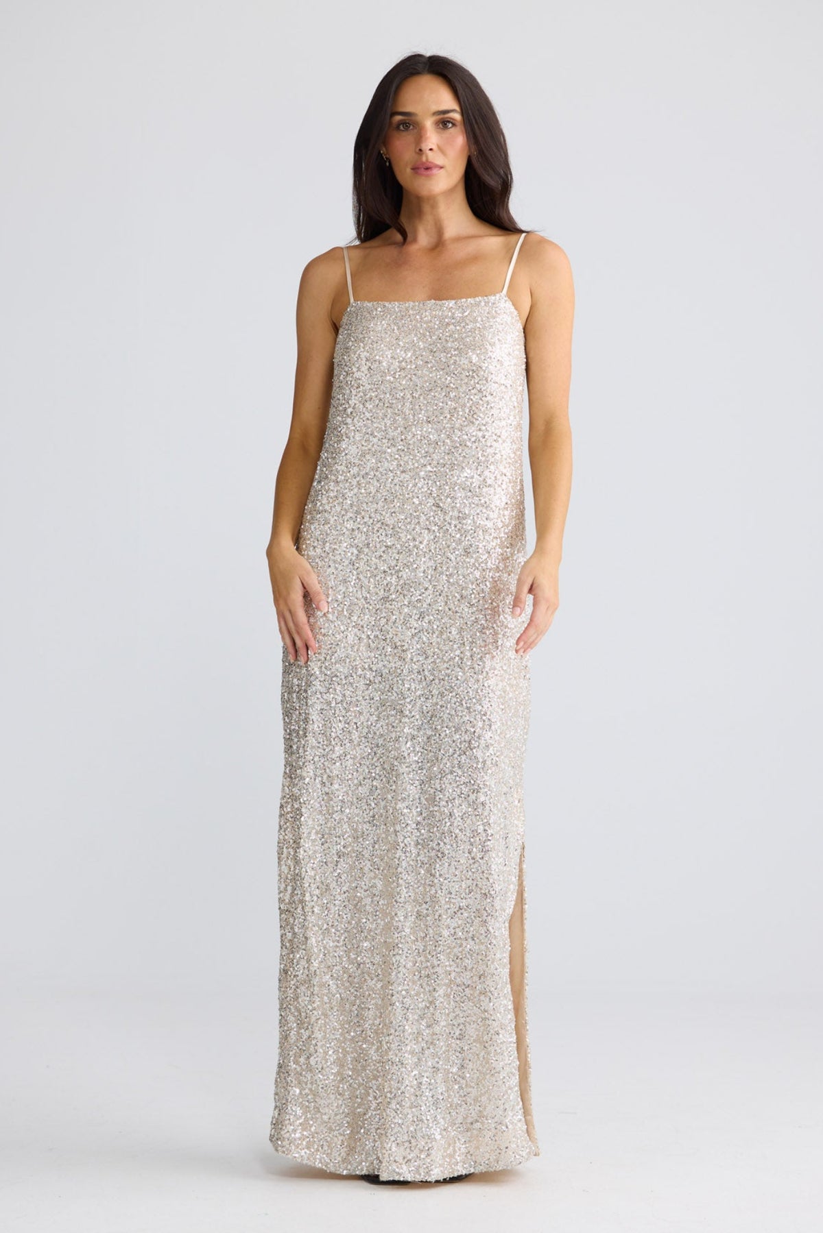 A woman modeling a floor-length champagne sequin slip dress with spaghetti straps and a thigh-high slit, photographed against a seamless light gray studio background by Brave + True.