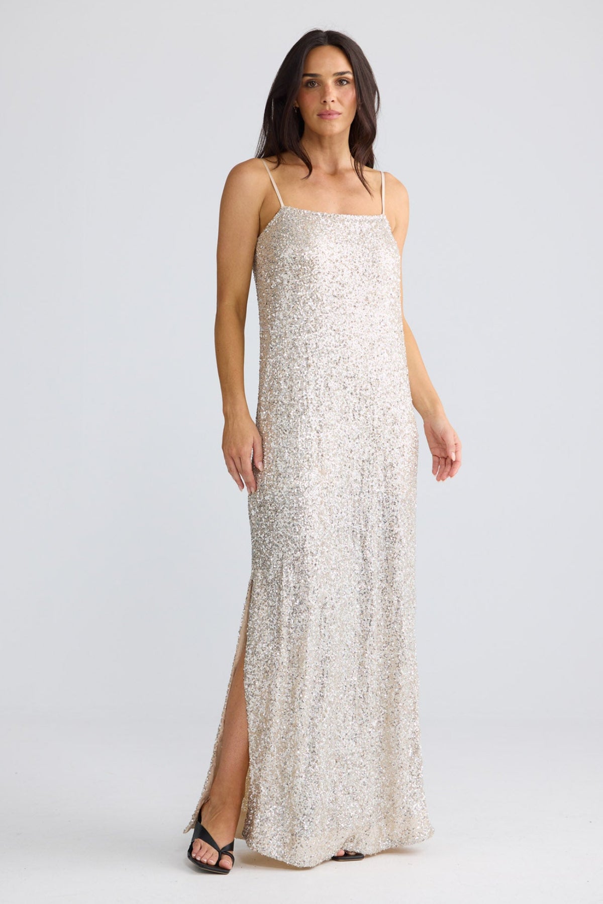 A woman modeling a sleeveless, floor-length champagne sequin gown with thin straps and a thigh-high slit, photographed against a light gray studio background by Brave + True.