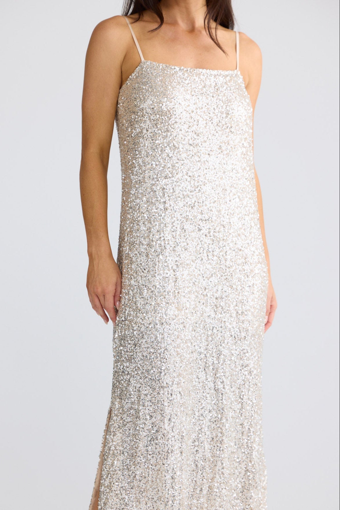 A woman wearing a floor-length champagne sequin slip dress with thin spaghetti straps and a thigh-high slit, photographed against a light gray studio background by Brave + True.