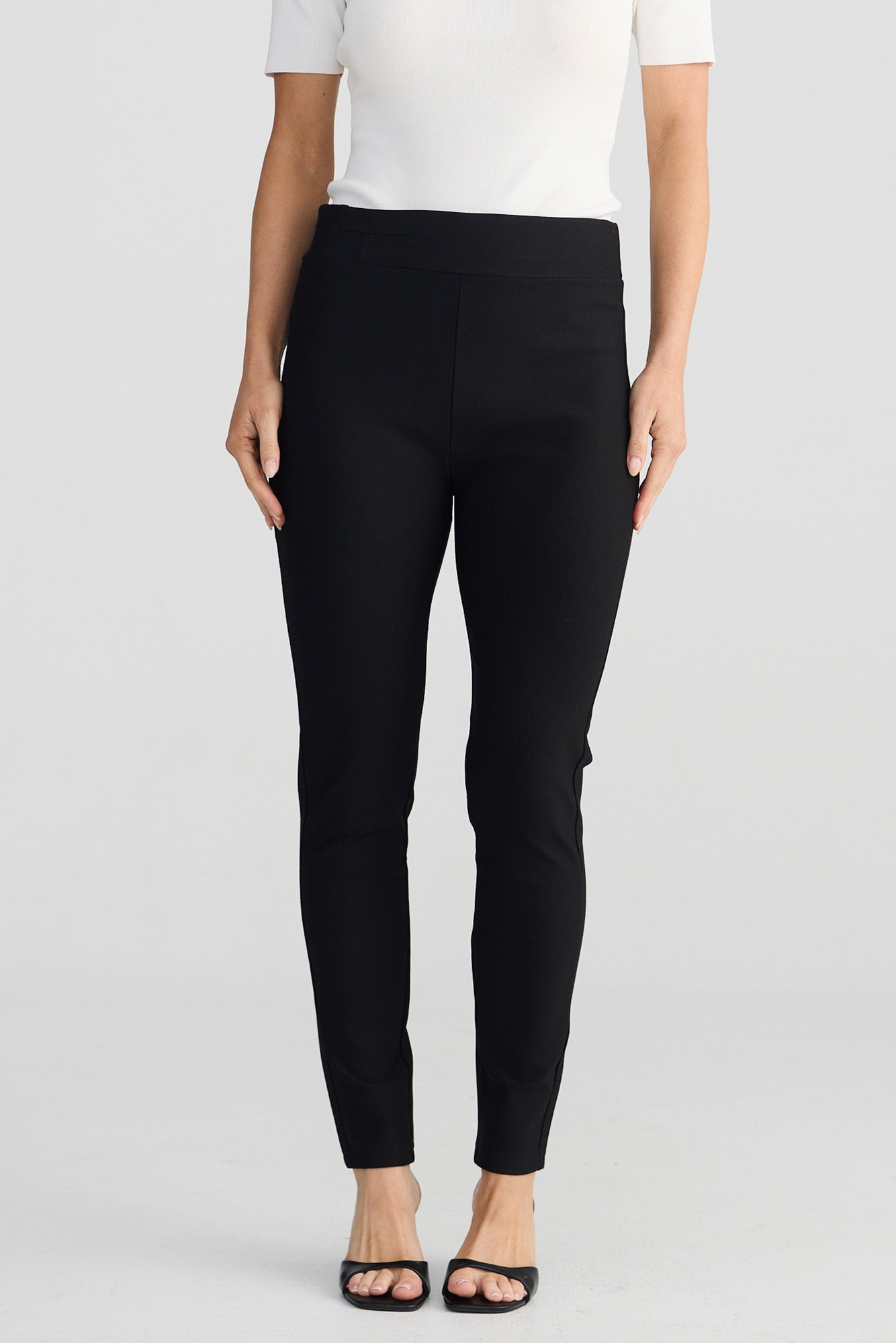 A model wearing black slim-fit ankle-length pants with a wide waistband, styled with a white top and black flat sandals against a light gray studio background.