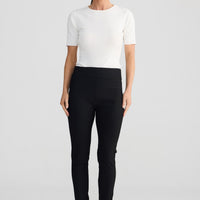 A woman stands against a light gray studio background wearing a fitted white short-sleeve top, black slim-fit pants, and black flat sandals.