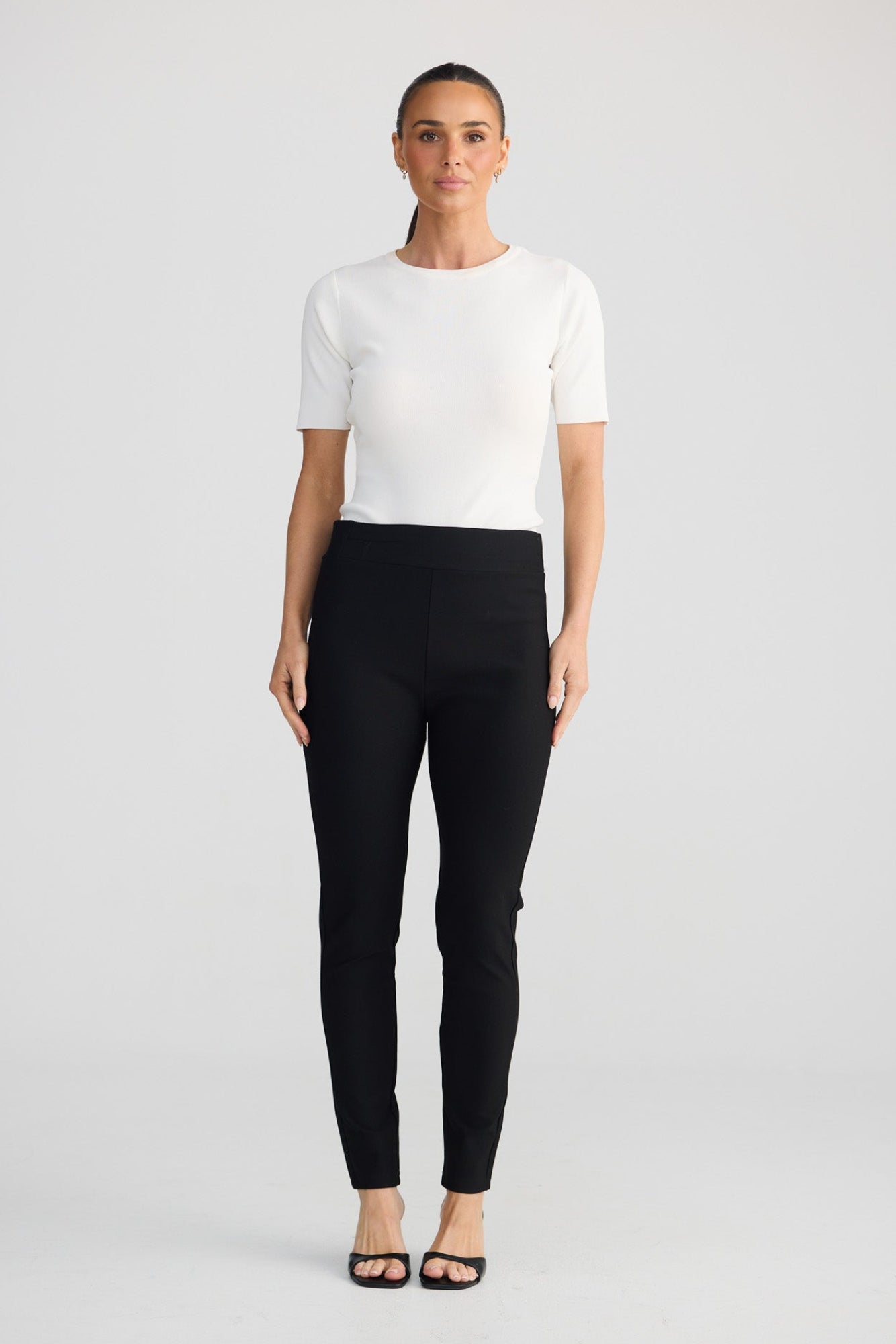 A woman stands against a light gray studio background wearing a fitted white short-sleeve top, black slim-fit pants, and black flat sandals.