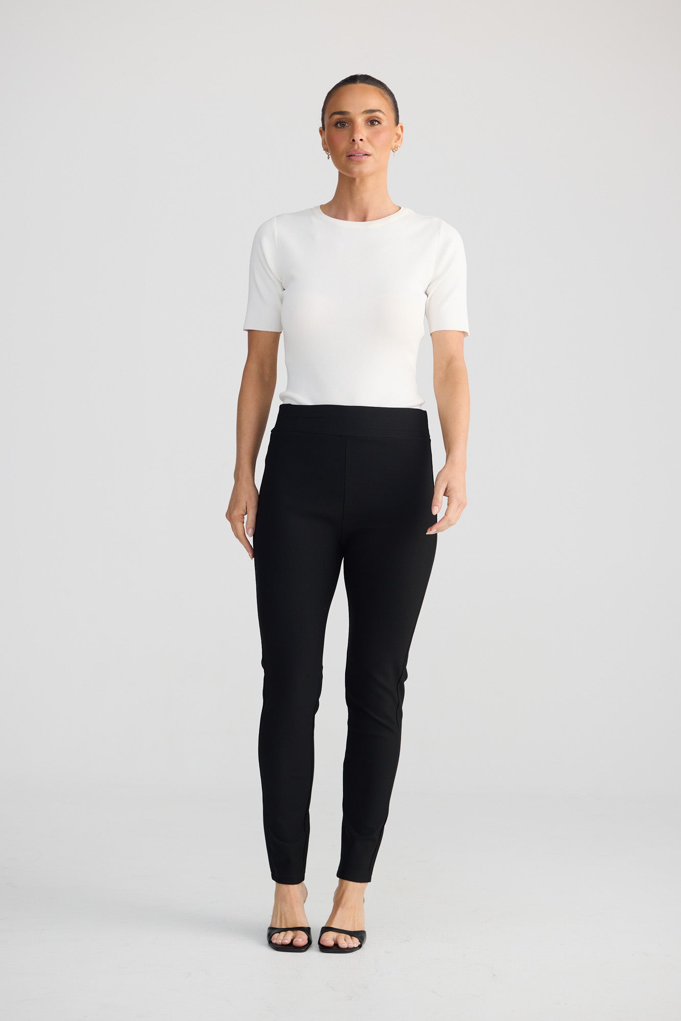 A woman stands against a light gray studio background wearing a fitted white short-sleeve top, black high-waisted slim-fit pants, and black flat sandals.