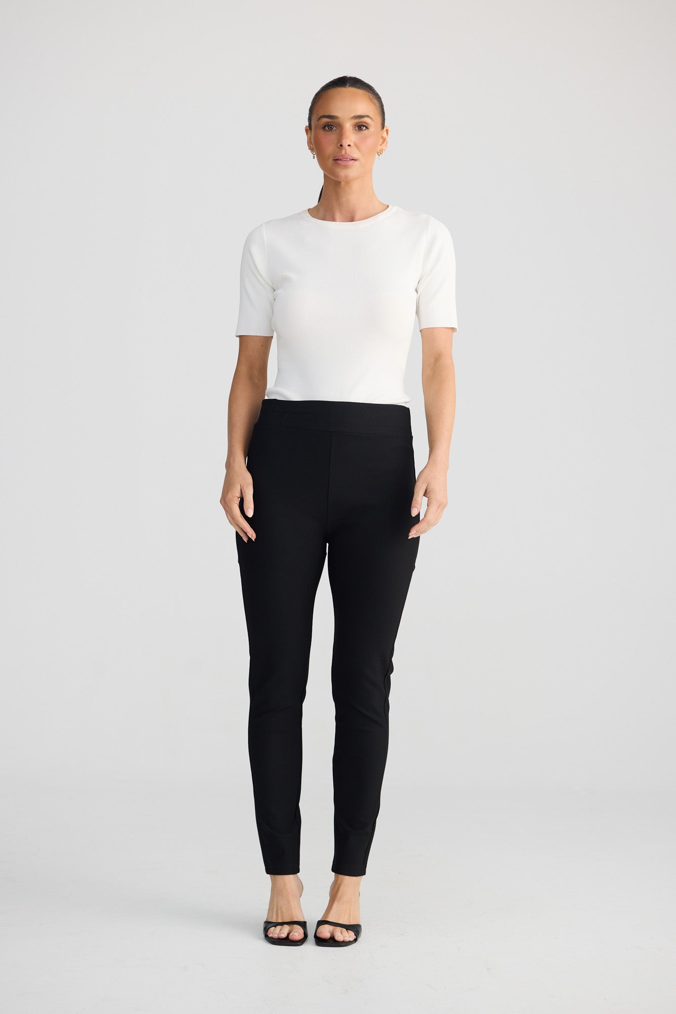 A woman stands against a light gray studio background wearing a fitted white short-sleeve top, black slim-fit pants, and black sandals.