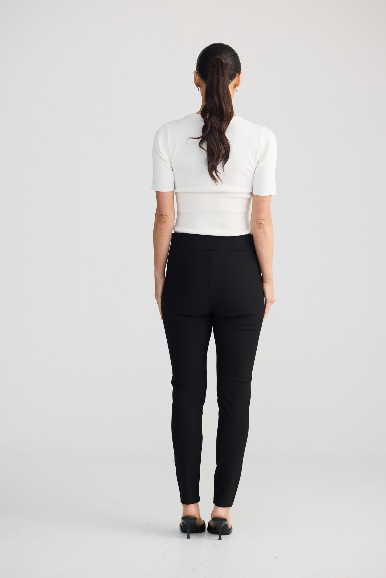 A woman viewed from behind wearing a fitted white short-sleeve top, black slim-fit pants, and black heels against a plain light gray studio background.