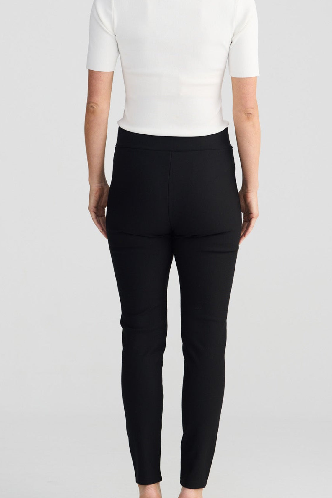 Rear view of a model wearing black slim-fit pants and a white short-sleeve top against a light gray studio background.