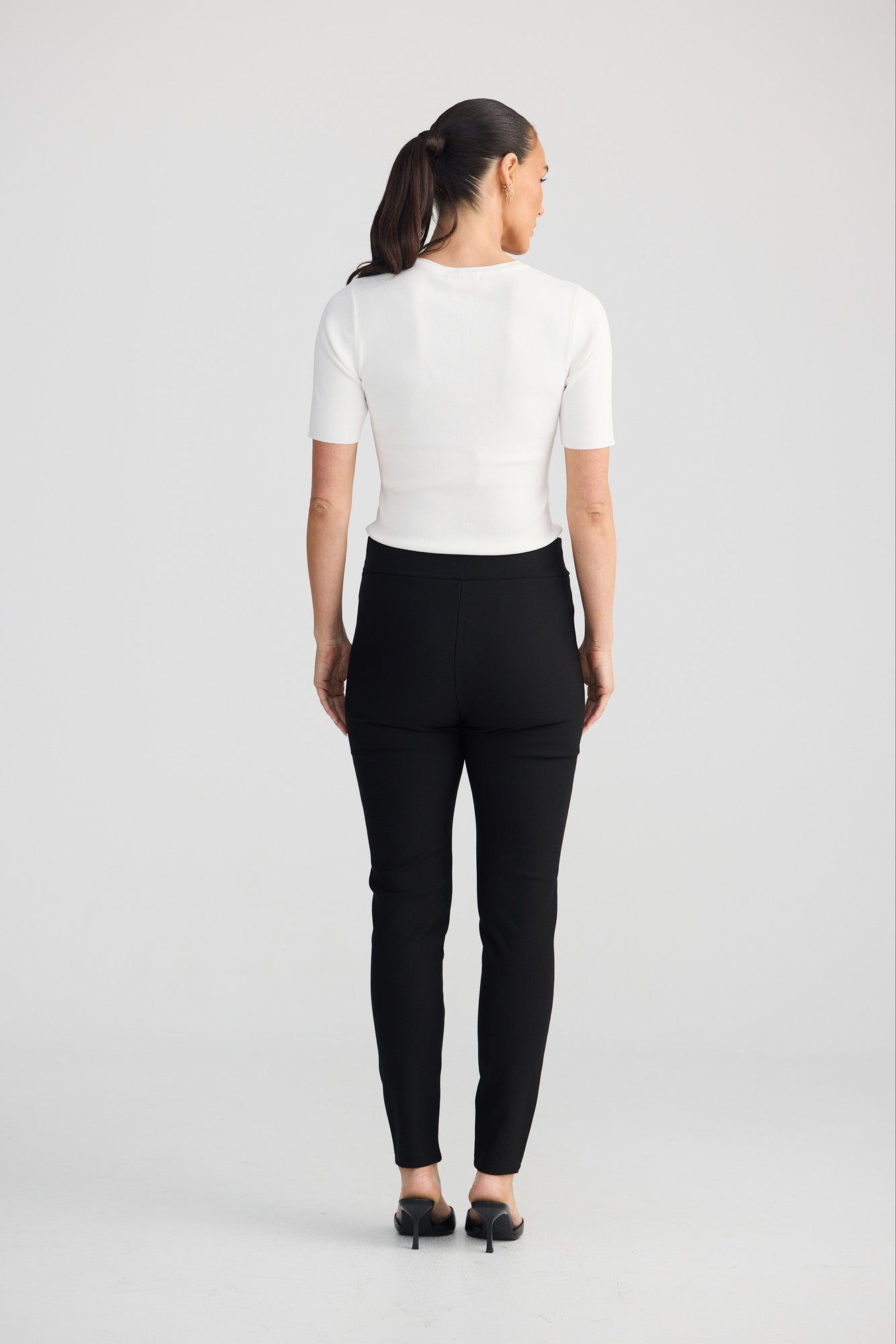 A woman viewed from the back wearing a fitted white short-sleeve top and black slim-fit pants, standing against a light gray studio background.