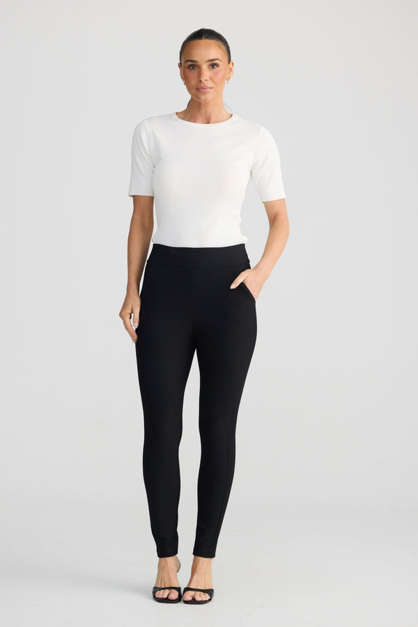 A woman modeling black high-waisted leggings with a white short-sleeve top, standing against a light gray studio background.