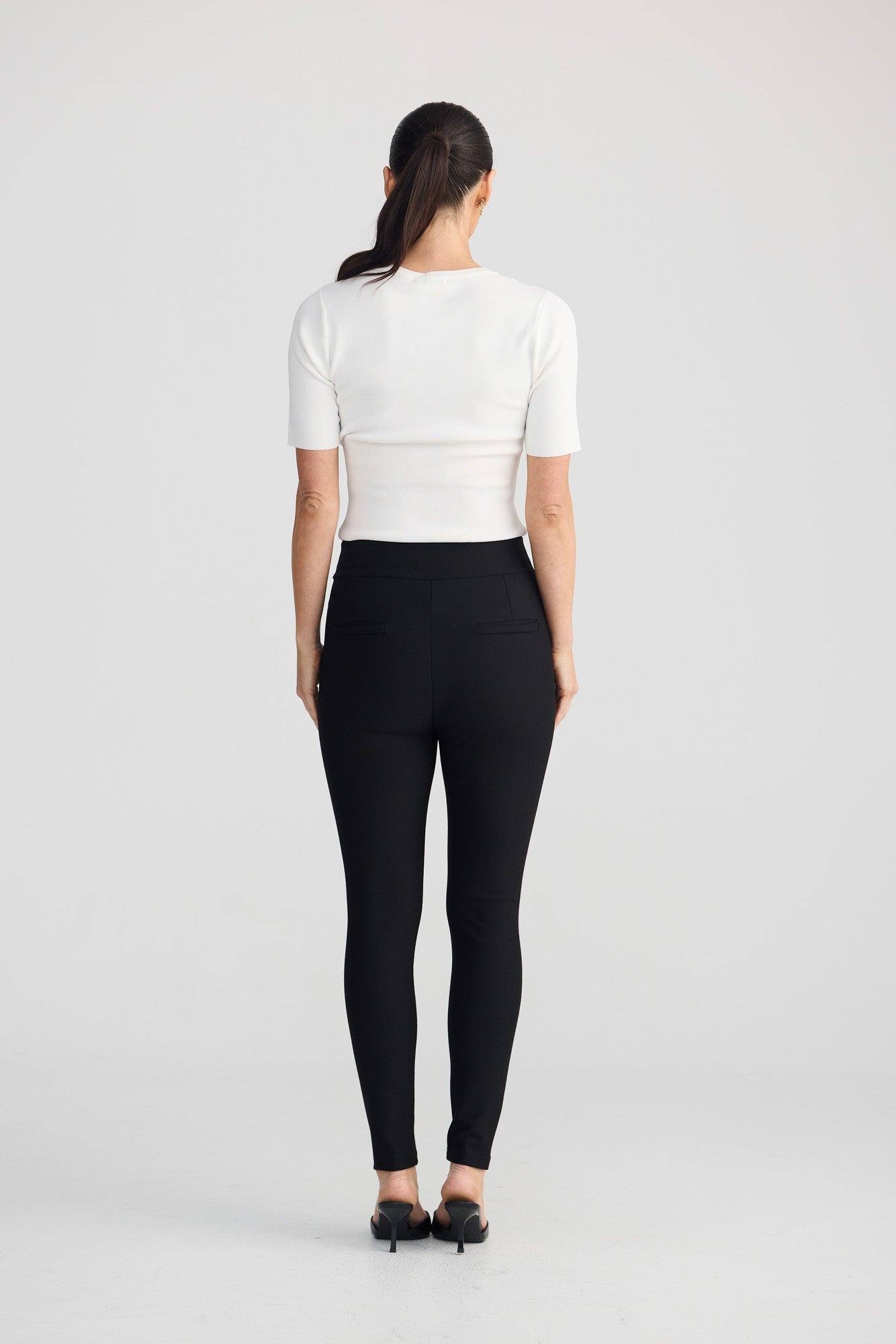 A woman viewed from the back wearing a fitted white short-sleeve top and black leggings, standing against a light gray studio background.