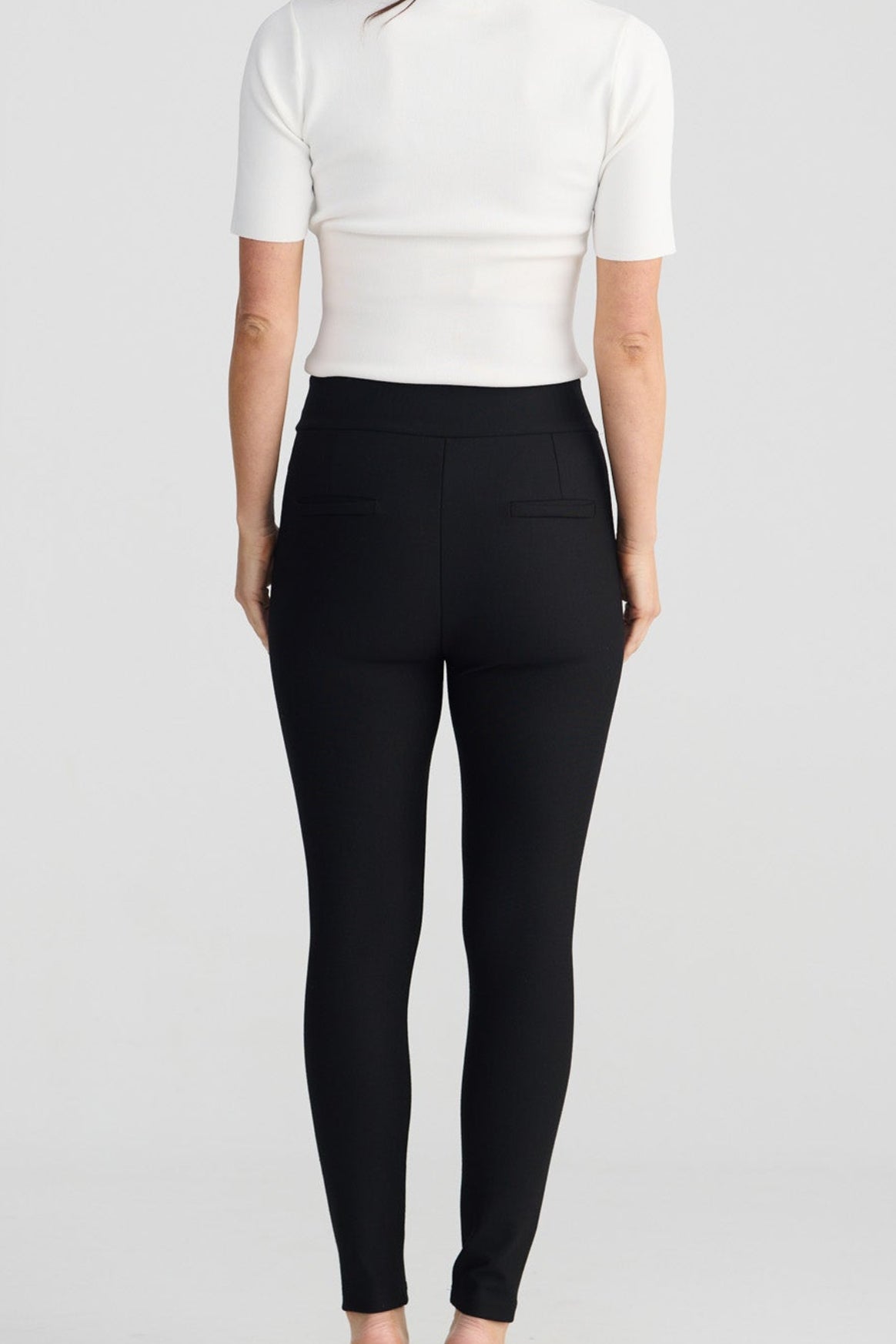 A woman viewed from behind wearing a fitted white short-sleeve top and black high-waisted leggings with black pointed flats, standing against a light gray studio background.
