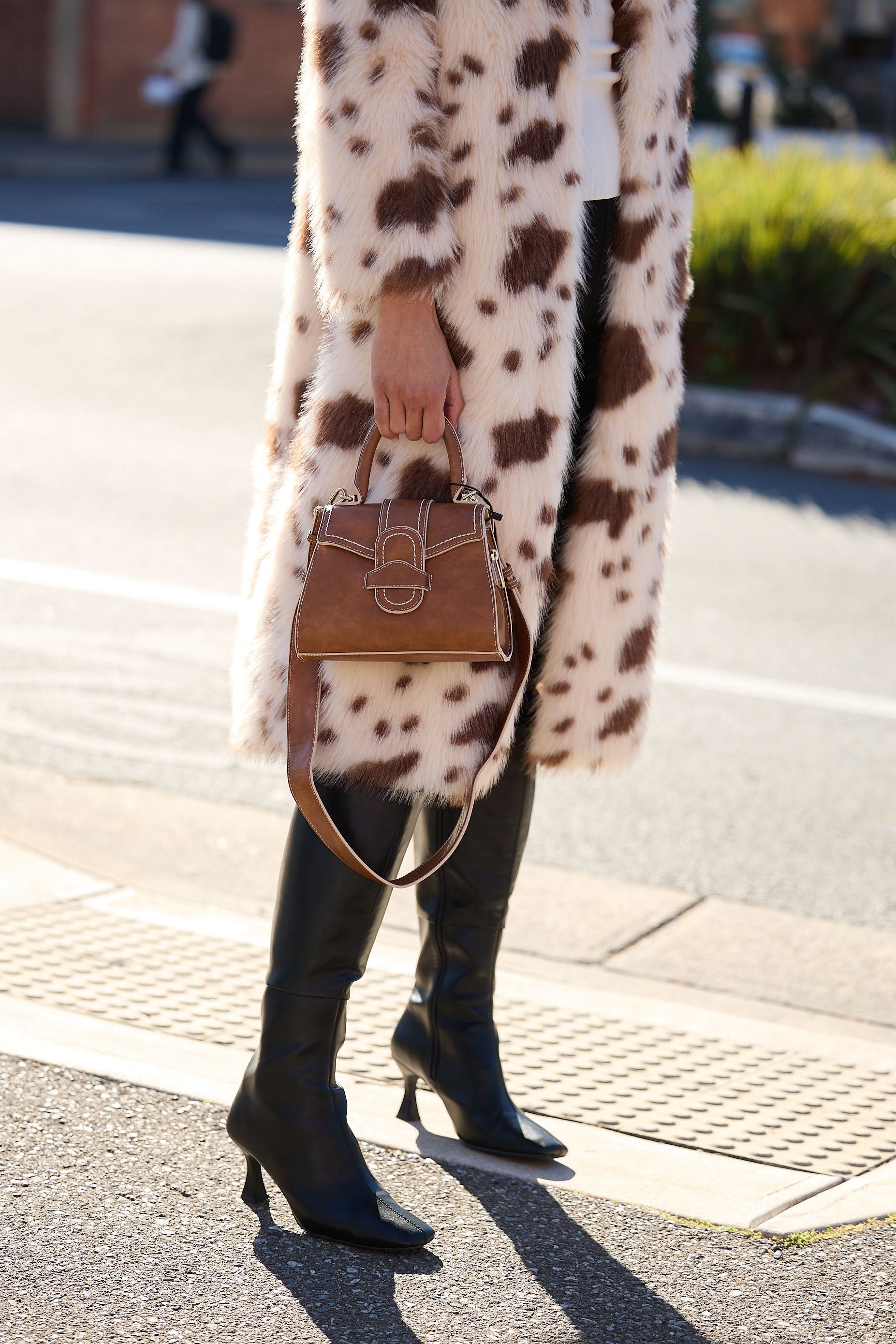 Person wearing a long coat with a pattern, holding a brown handbag on a street.