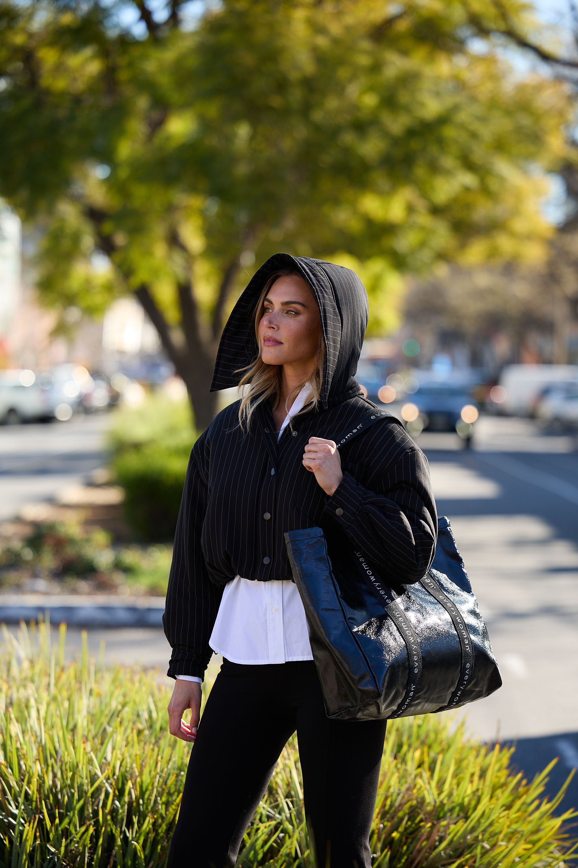 Woman walking outdoors wearing a black hoodie and carrying a large black bag.