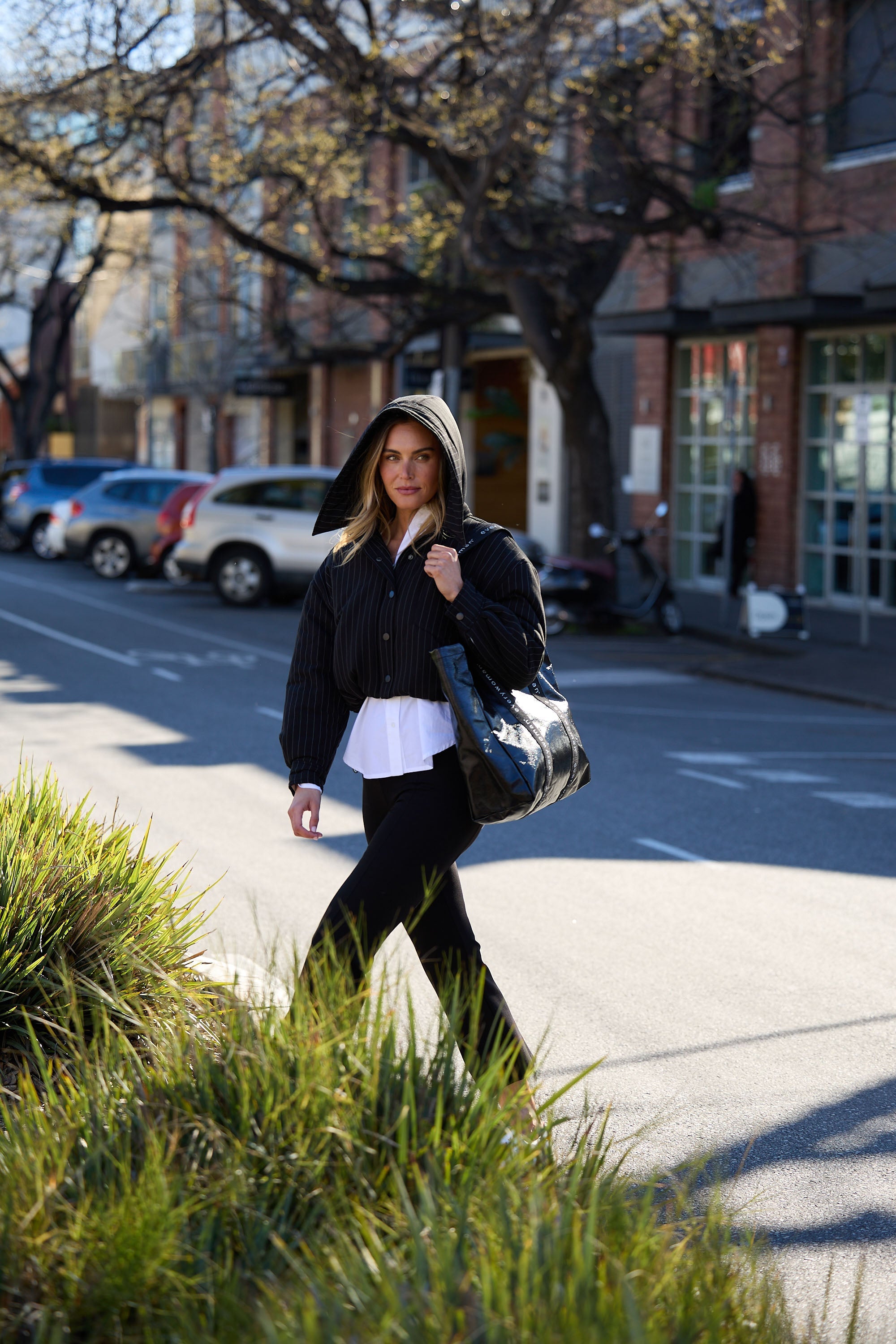Woman walking on a sidewalk holding a black handbag in an urban setting.