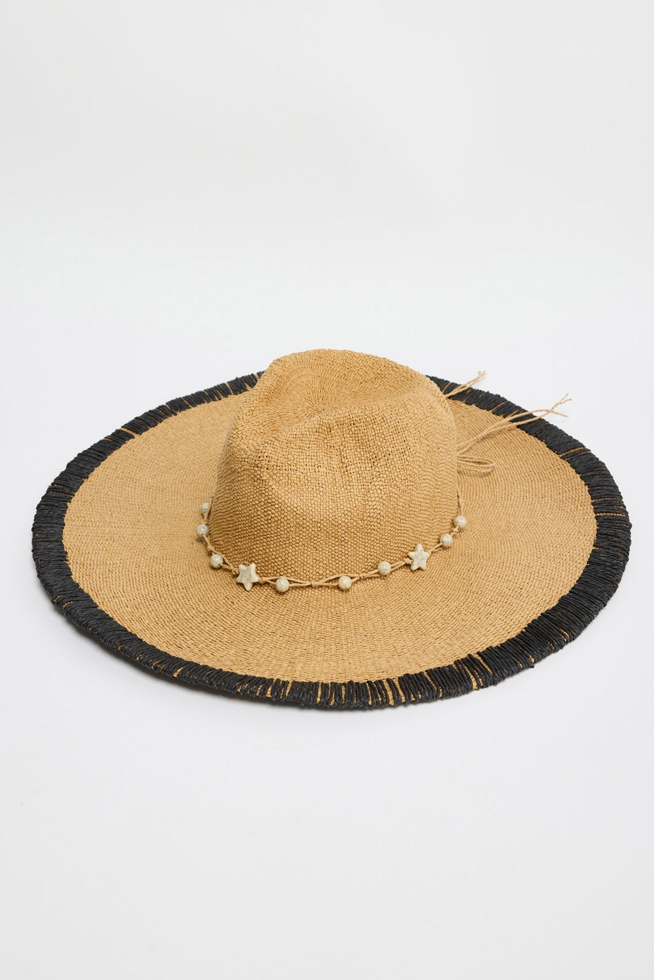 Carlitta Hat in natural woven straw with a black fringed brim edge, tan raffia tie and star and round bead detail, shown flat at an angle