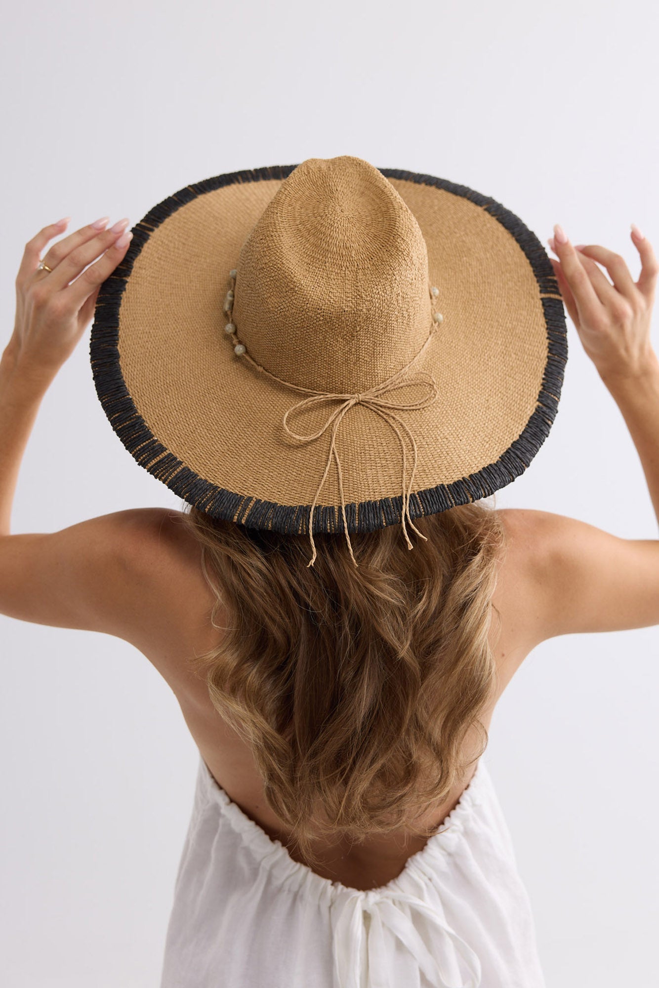 Woman wearing the Carlitta Hat from behind, holding the wide brim up over loose waves, paired with a white sundress