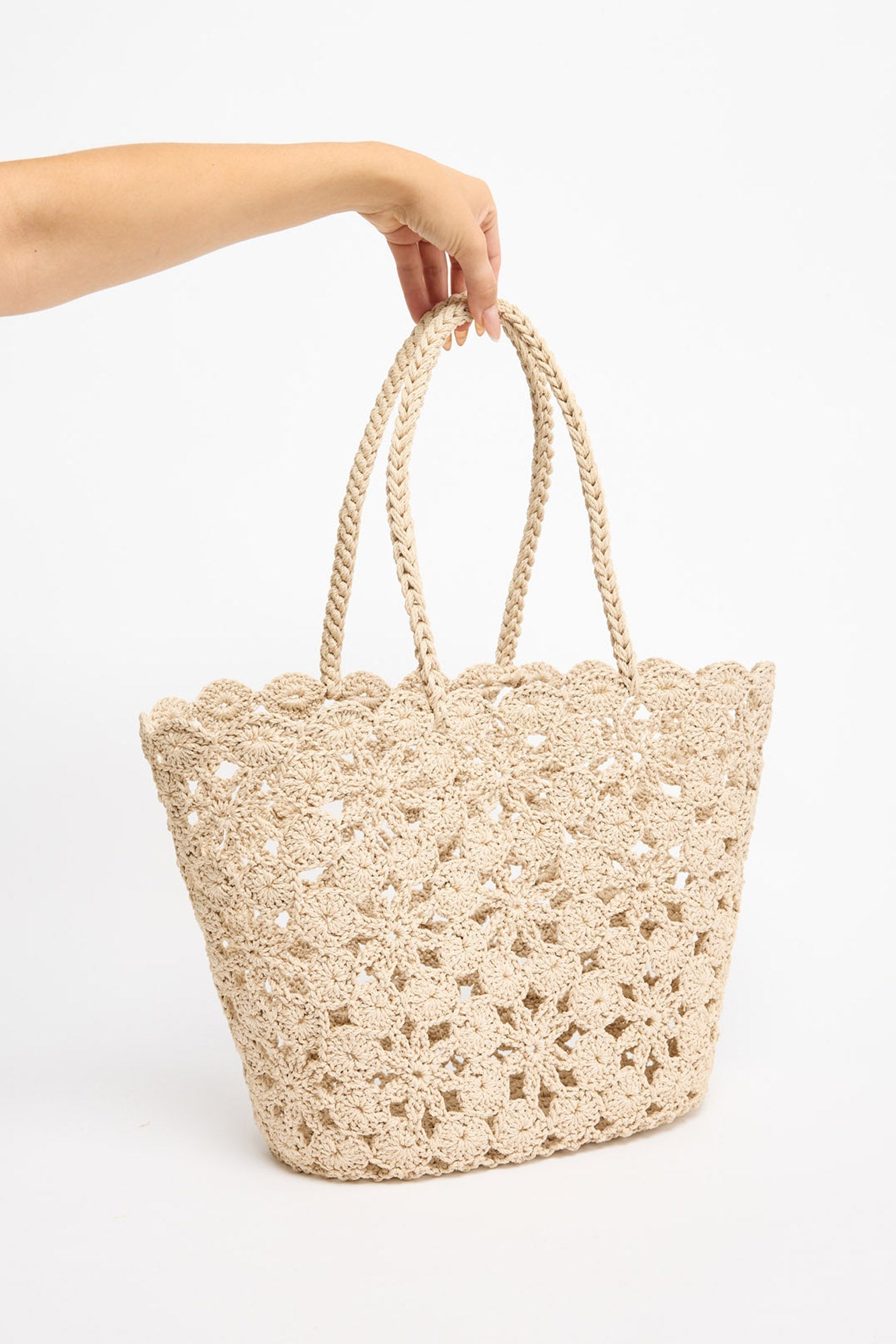 Large open-weave taupe crochet tote basket with twisted rope handles, shown held by hand against a white background