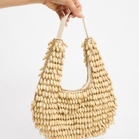Crescent-shaped shoulder bag fully covered in rows of natural cream oval wooden beads with gold-tone accent beads, held by hand against a white background