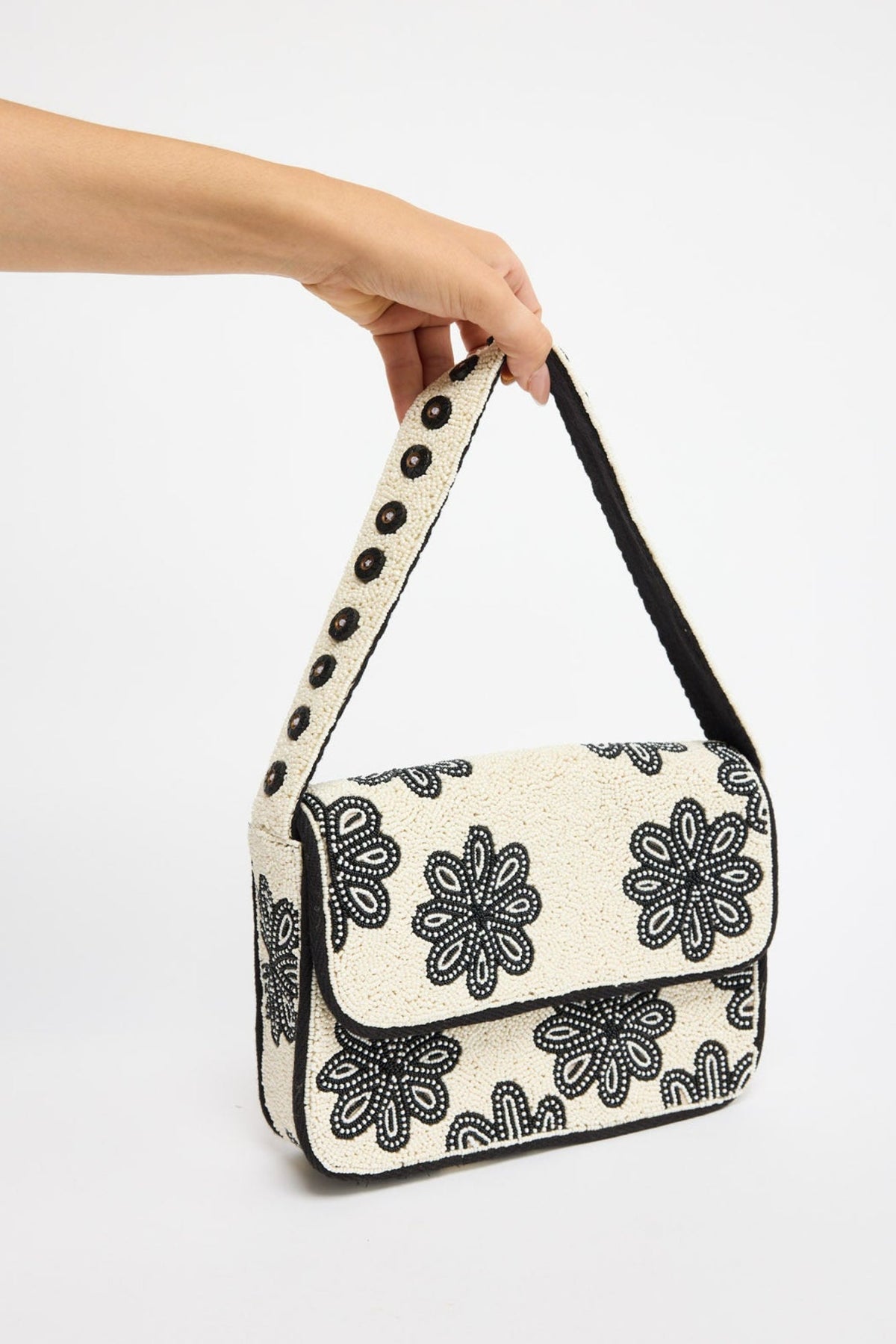 Structured rectangular shoulder bag fully covered in cream seed beads with bold black beaded floral motifs, black trim and a beaded strap with dark stud accents, held by hand against a white background