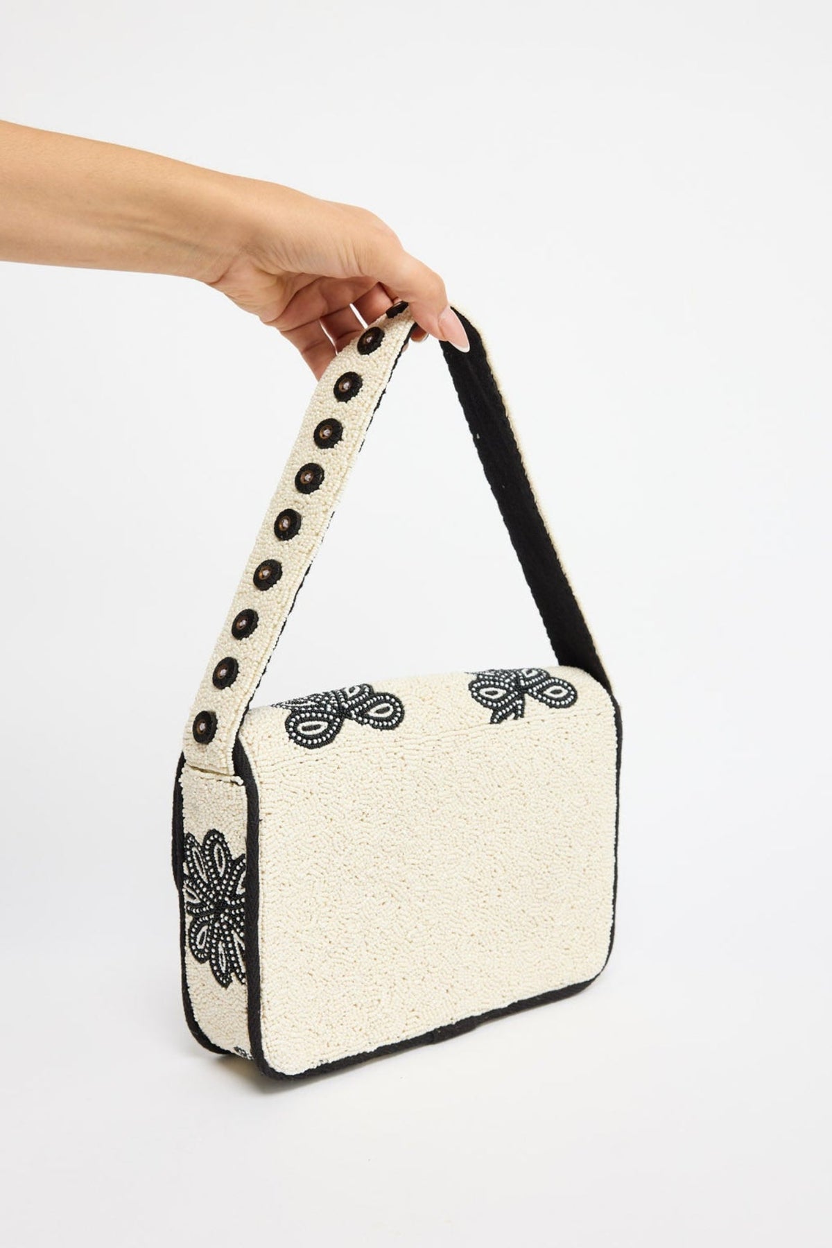 A hand holding a small cream-colored shoulder bag with black floral embroidery and a black strap, set against a plain white background.